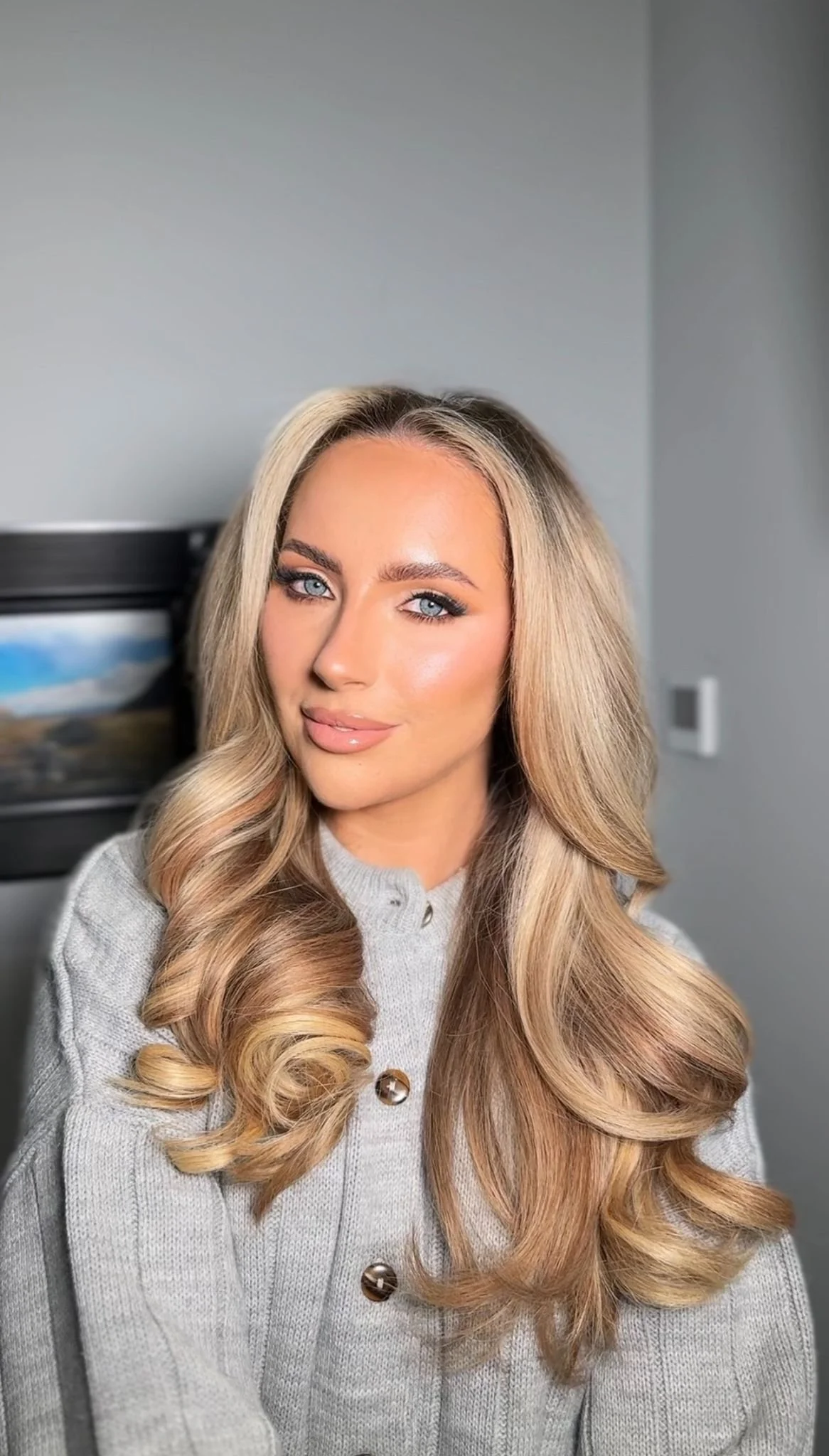 A woman with long, wavy blonde hair and blue eyes wearing a light gray button-up sweater, sitting indoors.