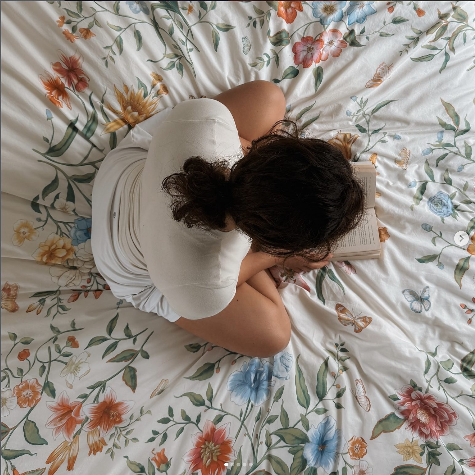 Person with dark curly hair sitting on a bed with floral bedding, reading a journaling.
