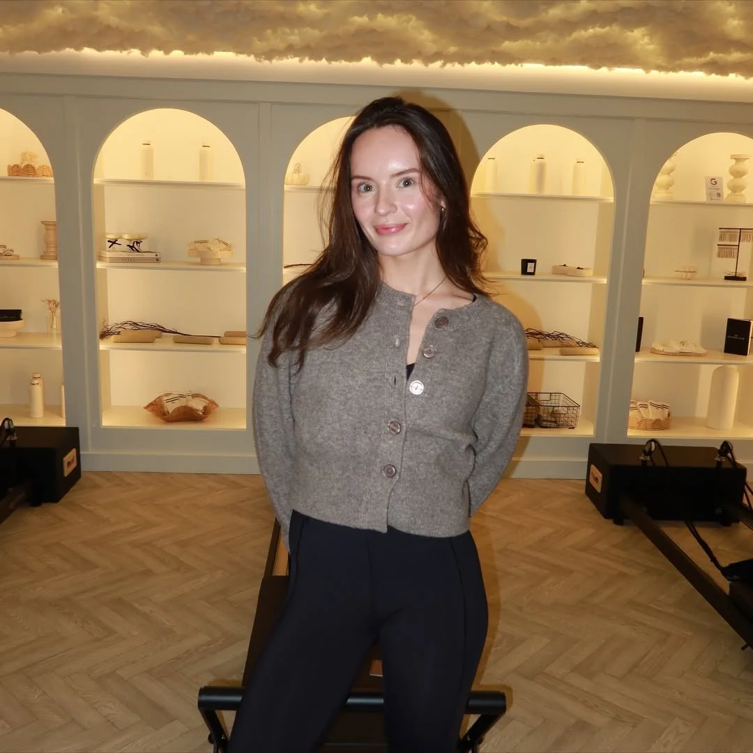 A woman with long dark hair wearing a gray cardigan and black pants standing indoors in front of white shelves with decorative items. The room has warm lighting and a wooden floor.