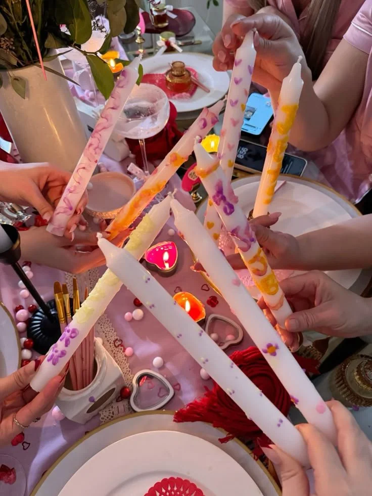People are painting candles, holding colourful twisted candles decorated with hearts and flowers, around a table with drinks, candles, and festive decorations.