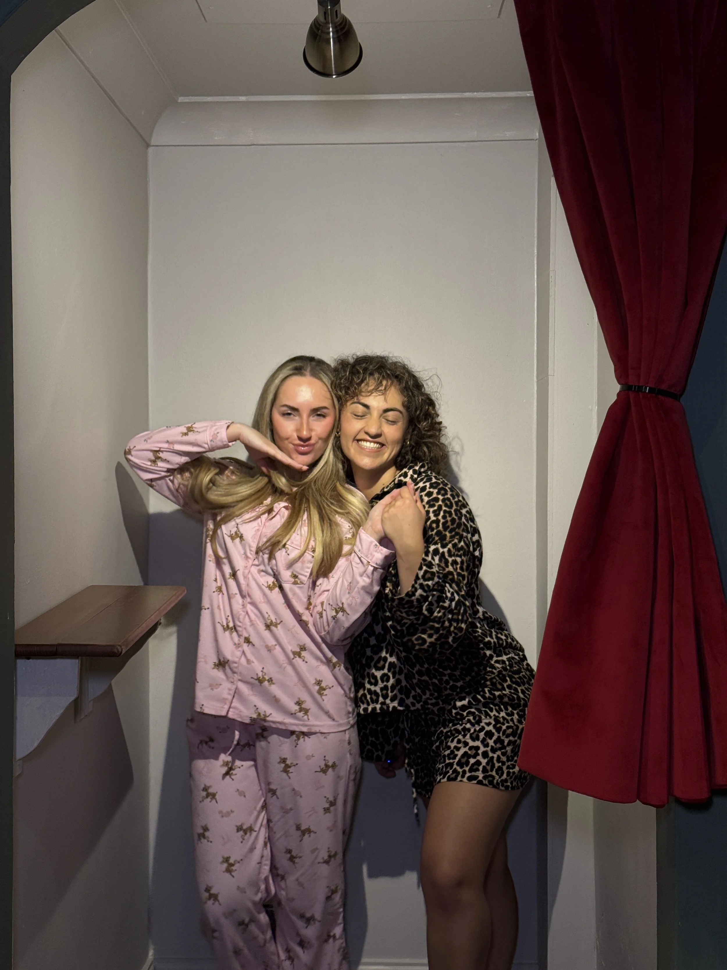 Two women in pajamas and leopard print outfit posing playfully in a small, curtained photo booth with a white wall background.