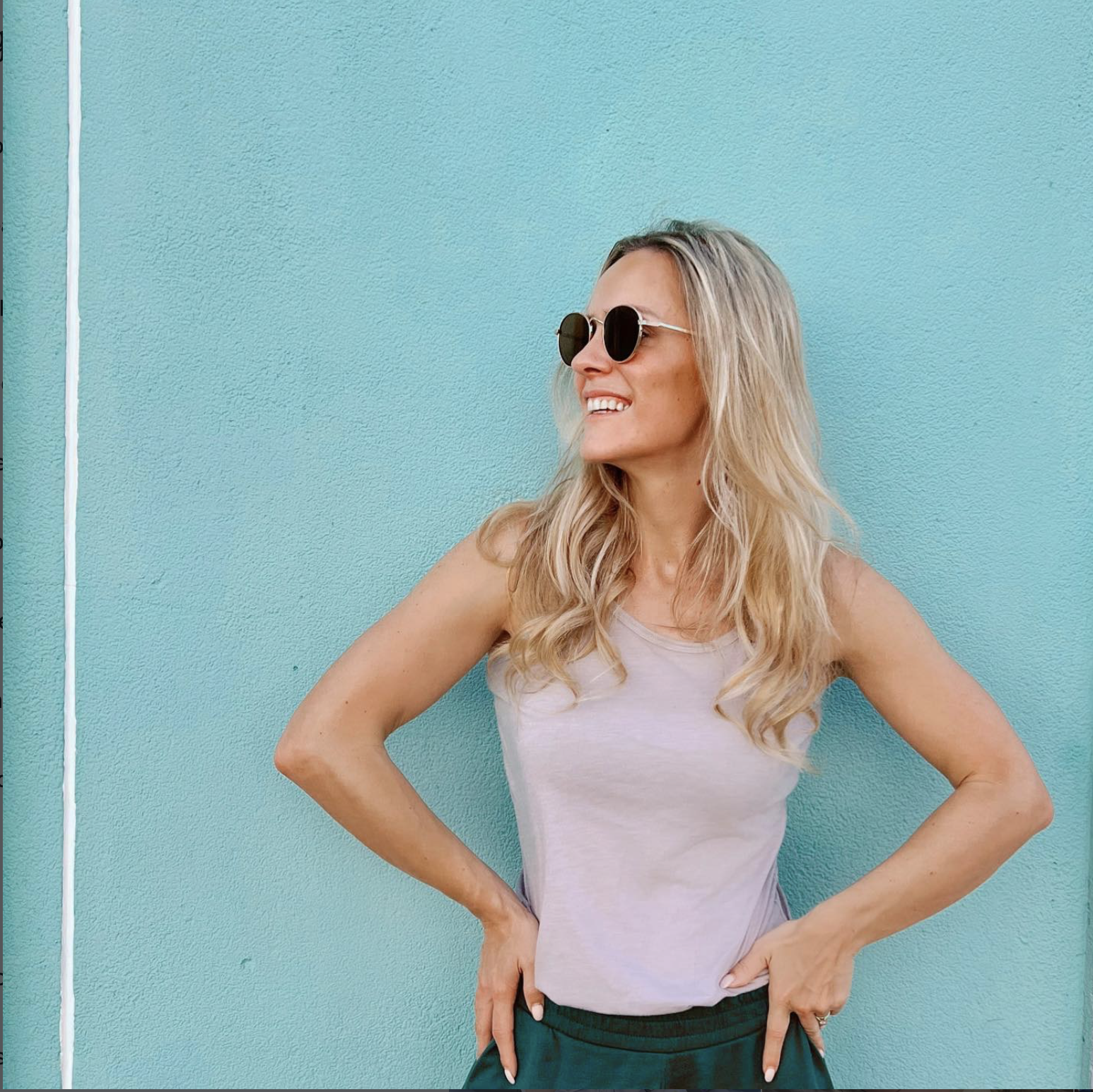 A woman with long blonde hair wearing sunglasses, a sleeveless beige top, and dark green pants standing against a bright blue wall, smiling and looking to her left.