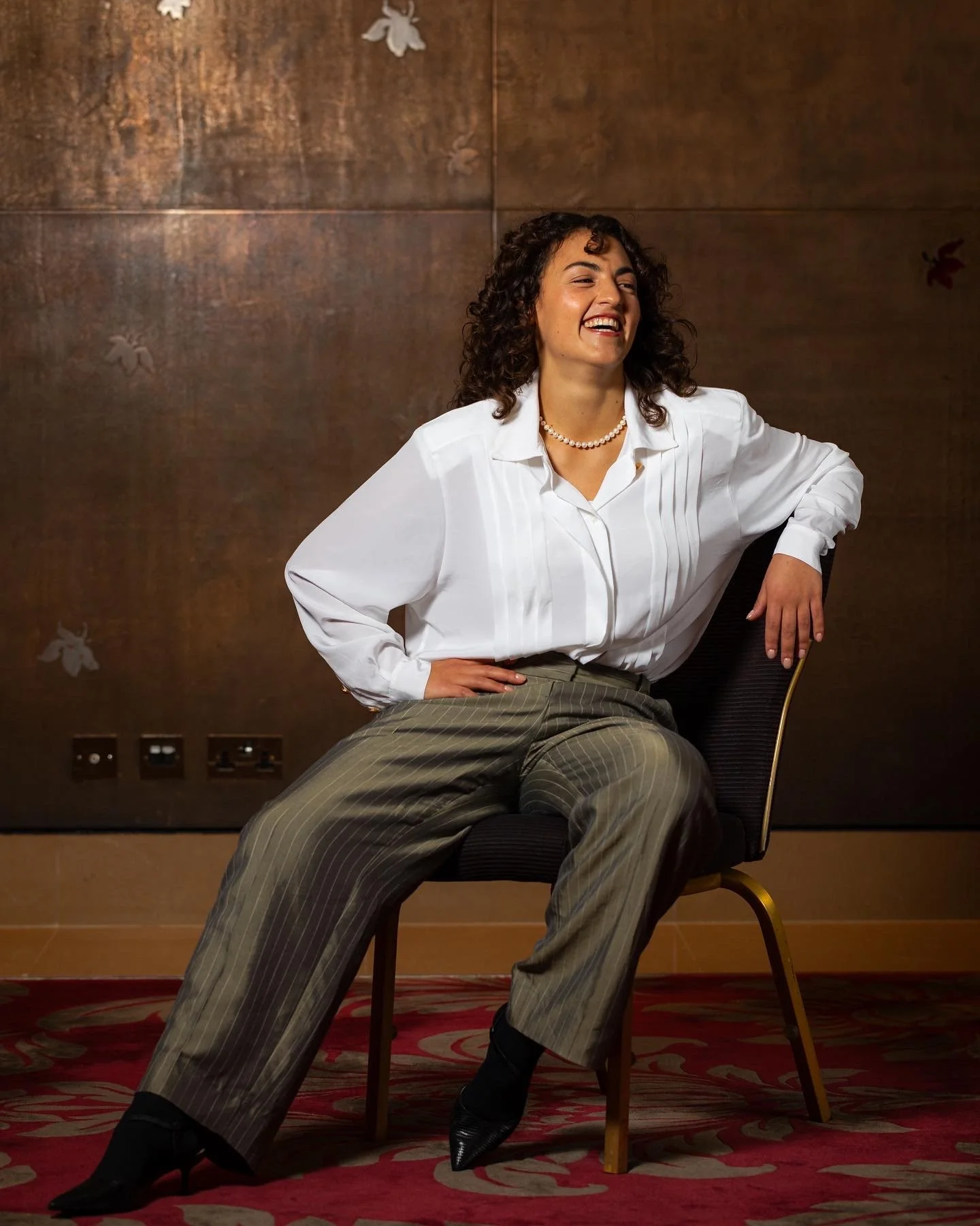 A woman with curly hair wearing a white blouse, striped pants, and black high-heeled shoes, sitting on a chair with a smile.
