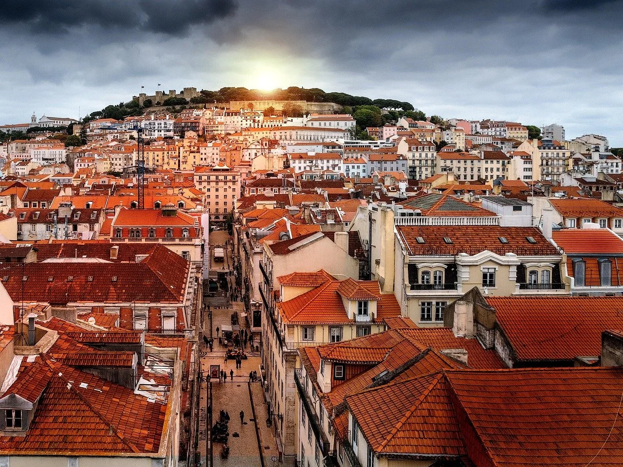 A cityscape with red-tiled roofs under a cloudy sky at sunset or sunrise, featuring a densely built area with many buildings extending up a hill.