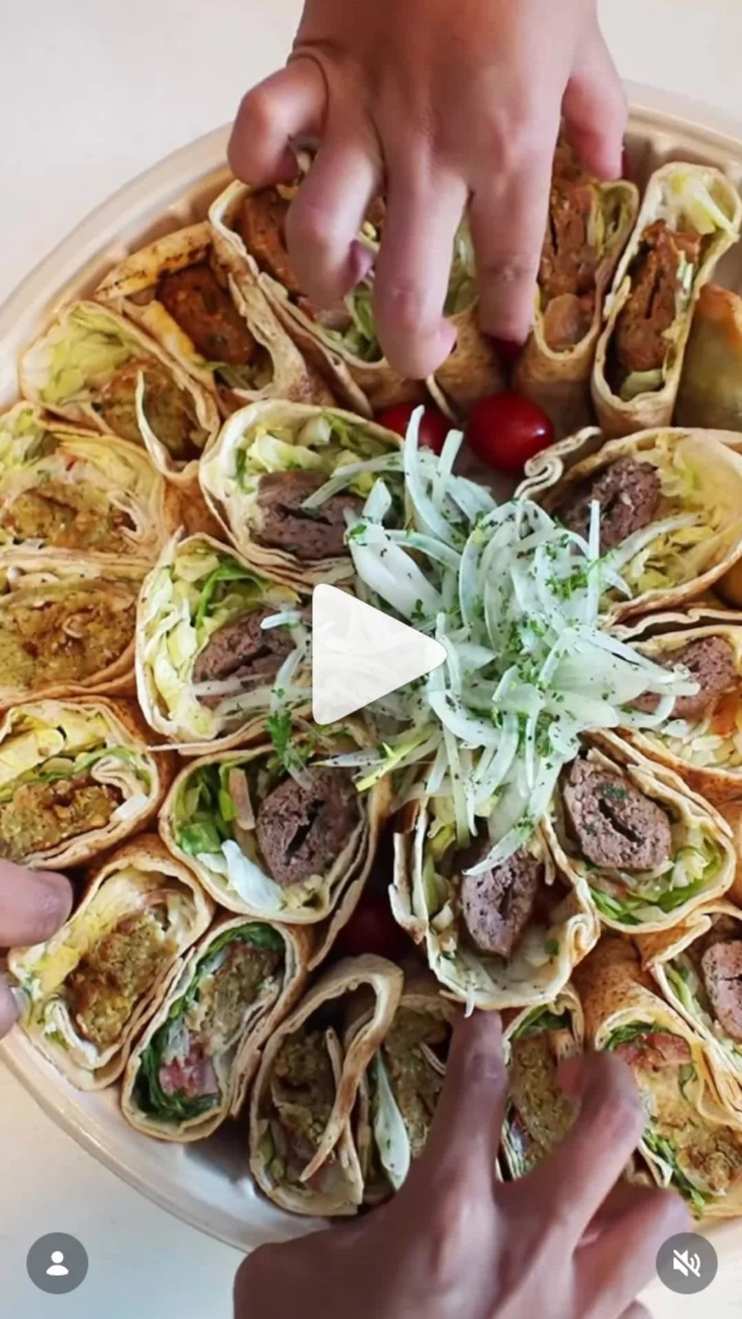 A large round tray filled with bite-sized wraps containing ground meat, lettuce, and other fillings. A hand is picking up a wrap, and there are cherry tomatoes and shredded cabbage in the center of the tray.