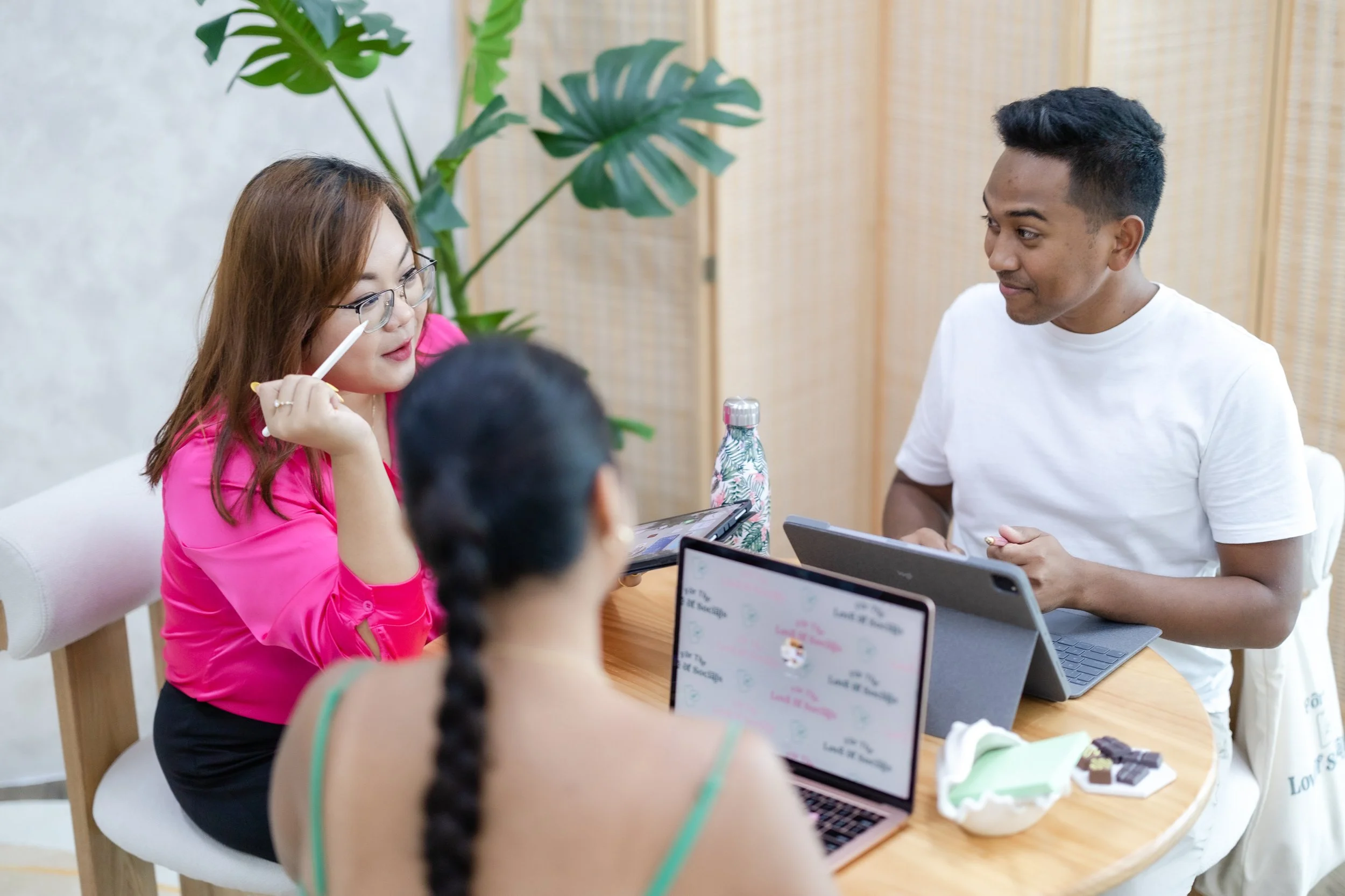 A group of people sitting around a table with laptops and drinks, engaging in conversation, with a green plant in the background.