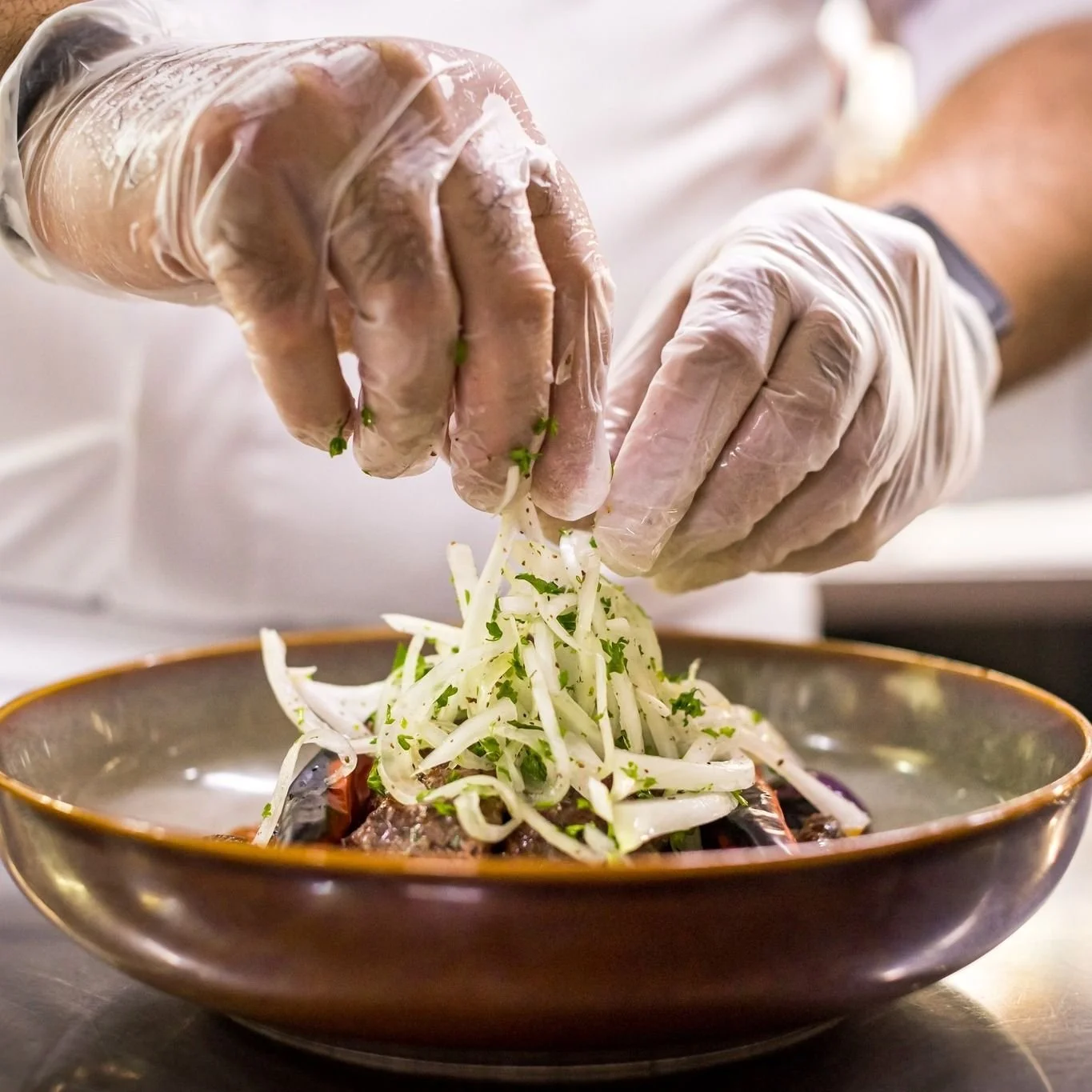 Person wearing gloves garnishing a bowl of Asian-inspired dish with shredded vegetables and herbs.