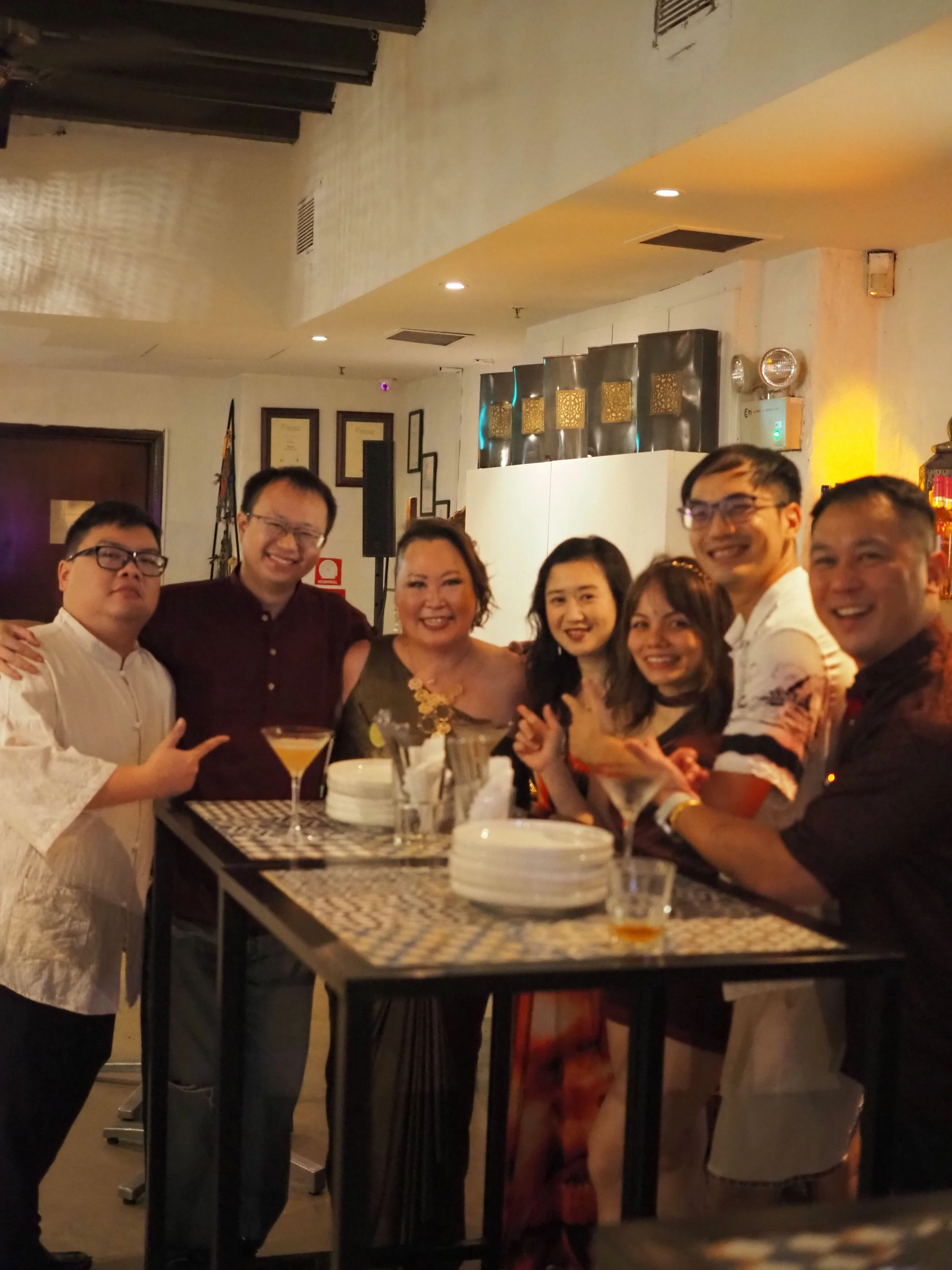 A group of smiling people gathered around a bar or counter in a restaurant or bar setting, with drinks and plates on the counter and framed pictures on the wall in the background.