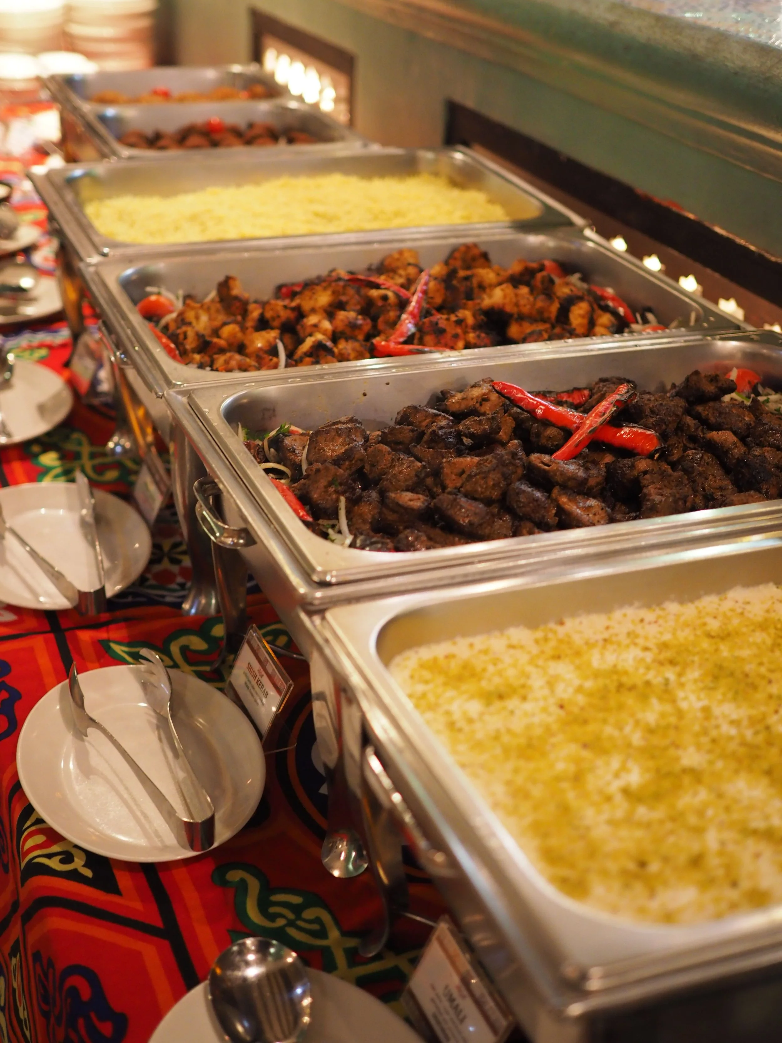 Buffet table with trays of food including yellow rice, grilled chicken, and beef, with plates, bowls, and utensils in front.
