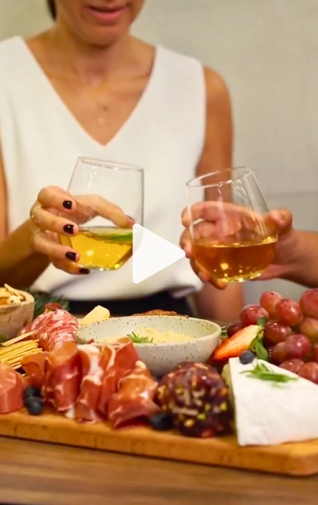 Two women clinking glasses of white wine or champagne over a table with a cheese and charcuterie platter, including grapes, sliced meats, cheese, and a bowl of dip.