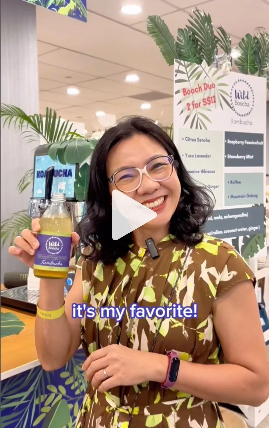 A woman smiling and holding a bottle of kombucha, with a sign behind her advertising various flavors and a promotion for two for $12. She is wearing glasses, a patterned dress, and a fitness tracker on her wrist, in a setting decorated with tropical 