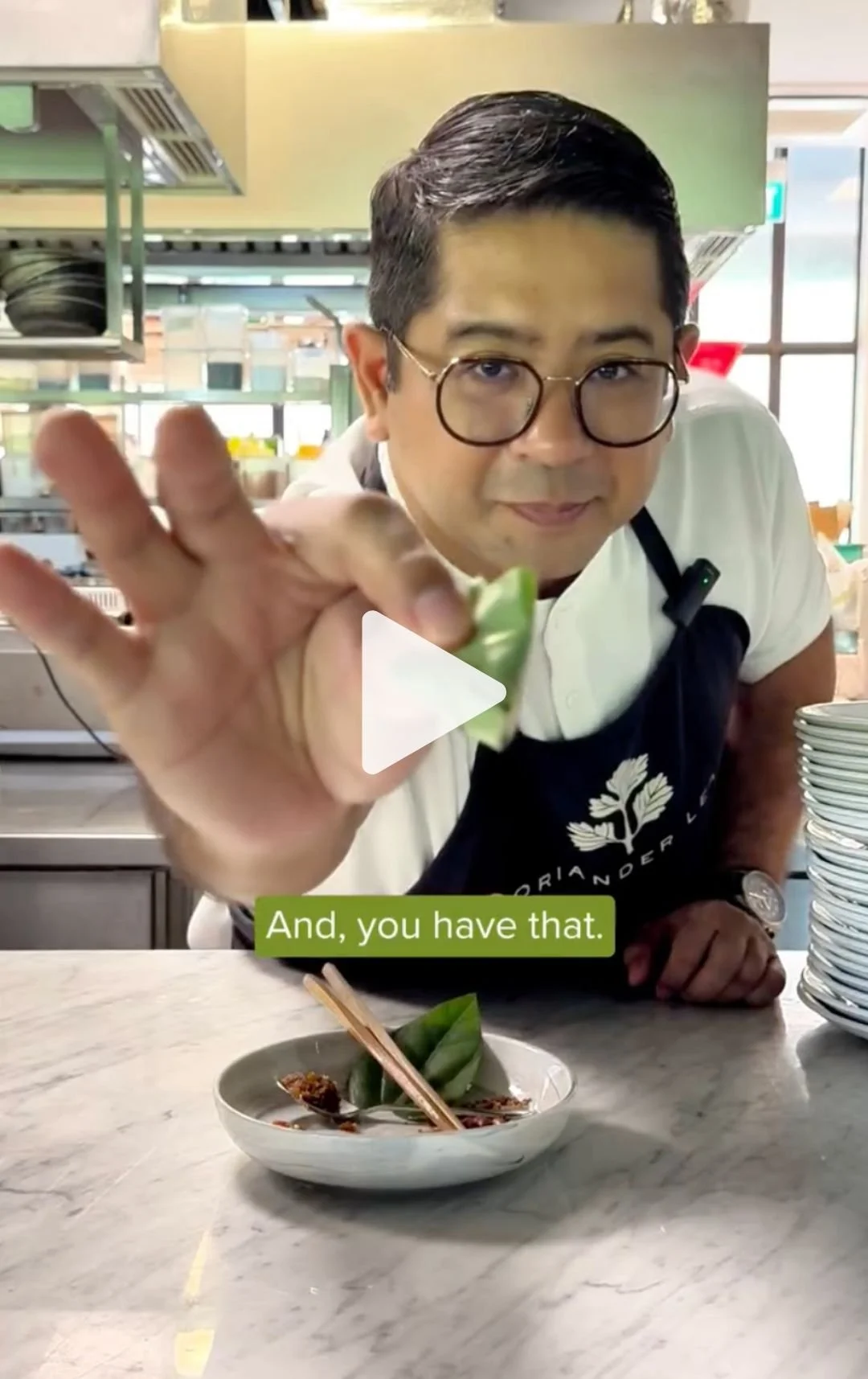 A man with glasses and a white shirt, wearing a black apron with a tree logo, is leaning over a kitchen counter and holding a piece of food towards the camera. The background shows a commercial kitchen with shelves and kitchen equipment. On the count