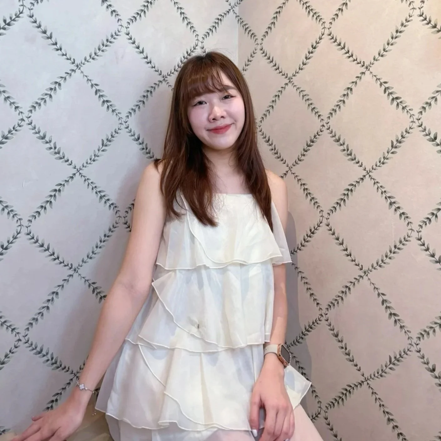 A young woman with shoulder-length brown hair and bangs, smiling, wearing a white, ruffled sleeveless dress, standing against a wall with a black leafy vine pattern.