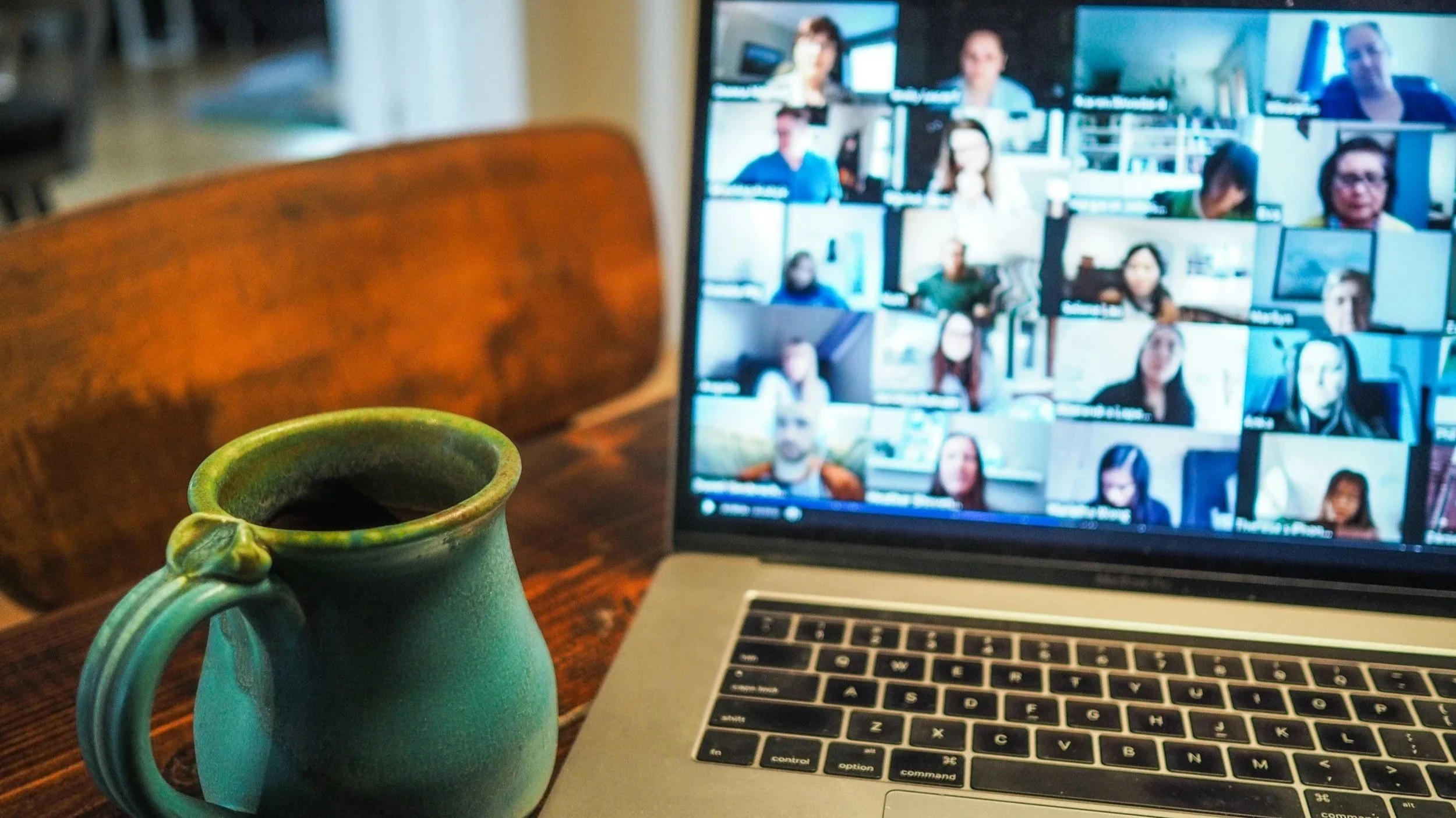 A teal ceramic mug and a silver laptop with a video call open showing multiple people, on a wooden table in a cozy indoor setting.
