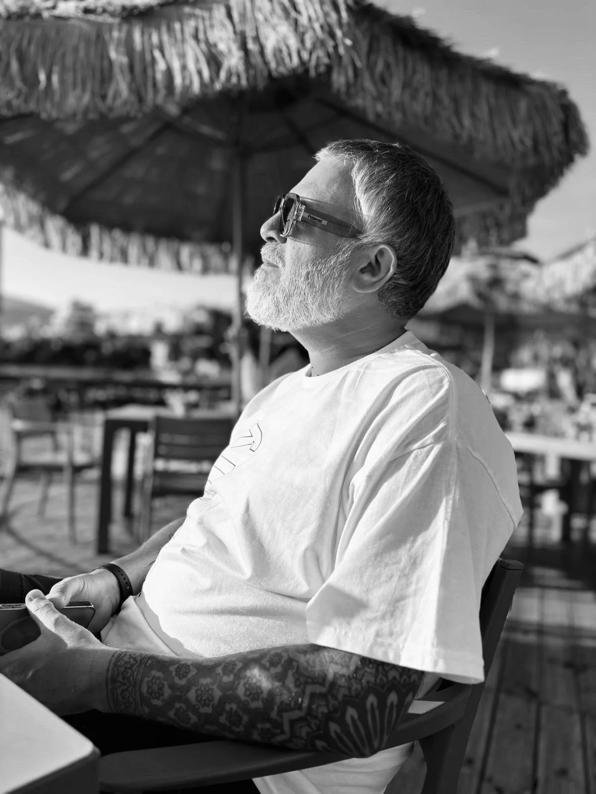 A man with a gray beard and sunglasses, wearing a white t-shirt and a tattooed arm, sitting outside under a large straw umbrella, holding a smartphone in his left hand.