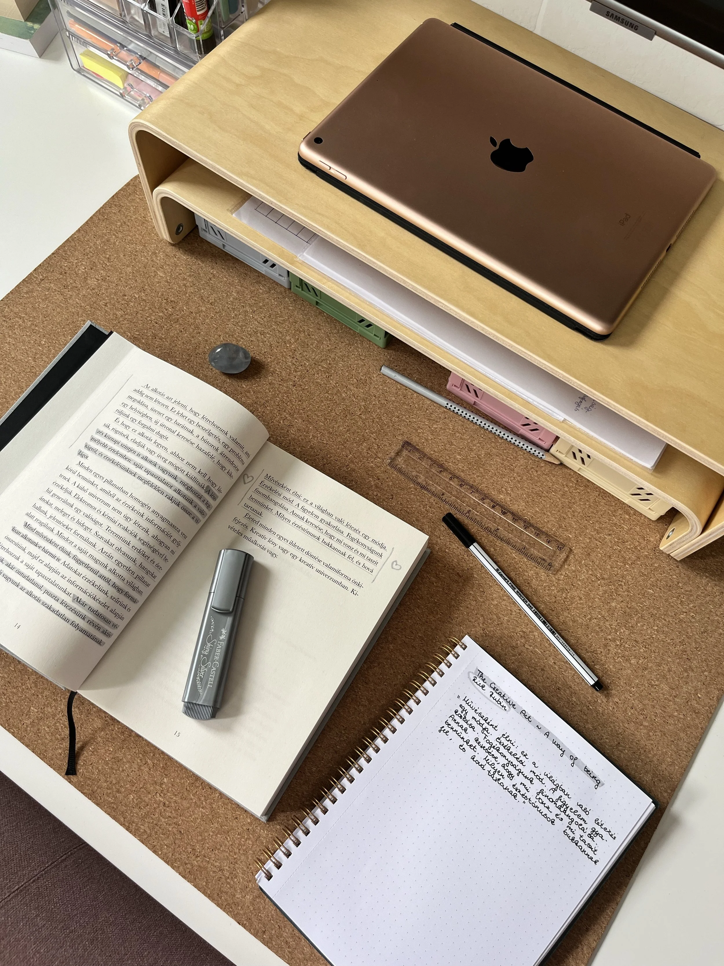 a notebook, book, and laptop with some writing tools lay on a corkboard desk
