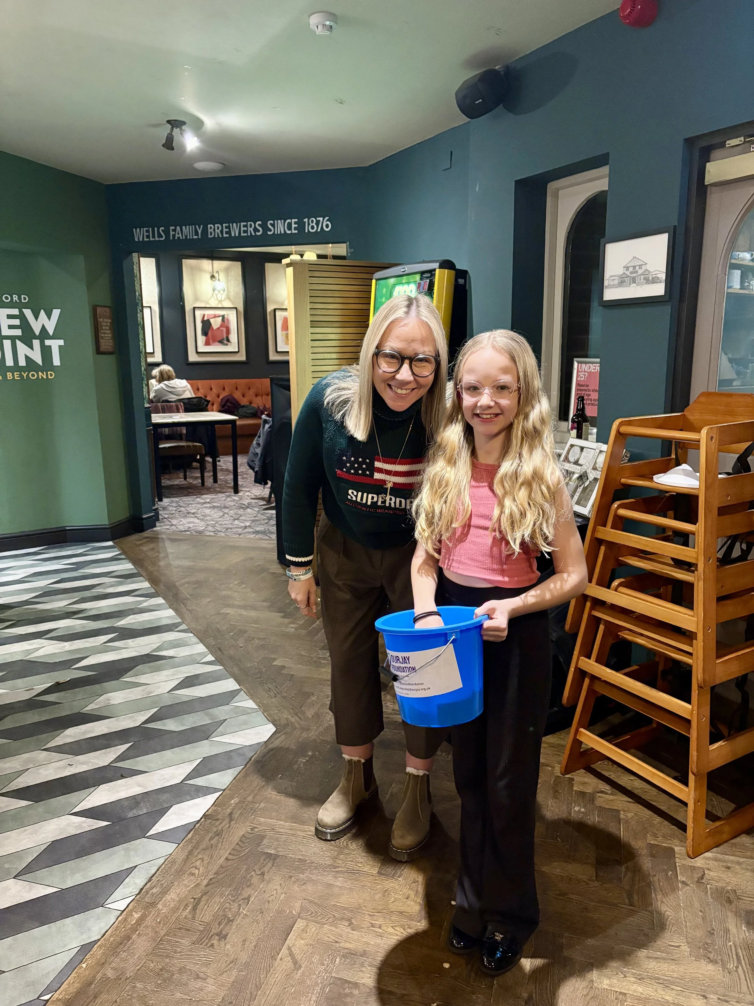 A woman and a young girl smiling and posing for a photo inside a restaurant or cafe, with the girl holding a blue donation bucket for a charity.