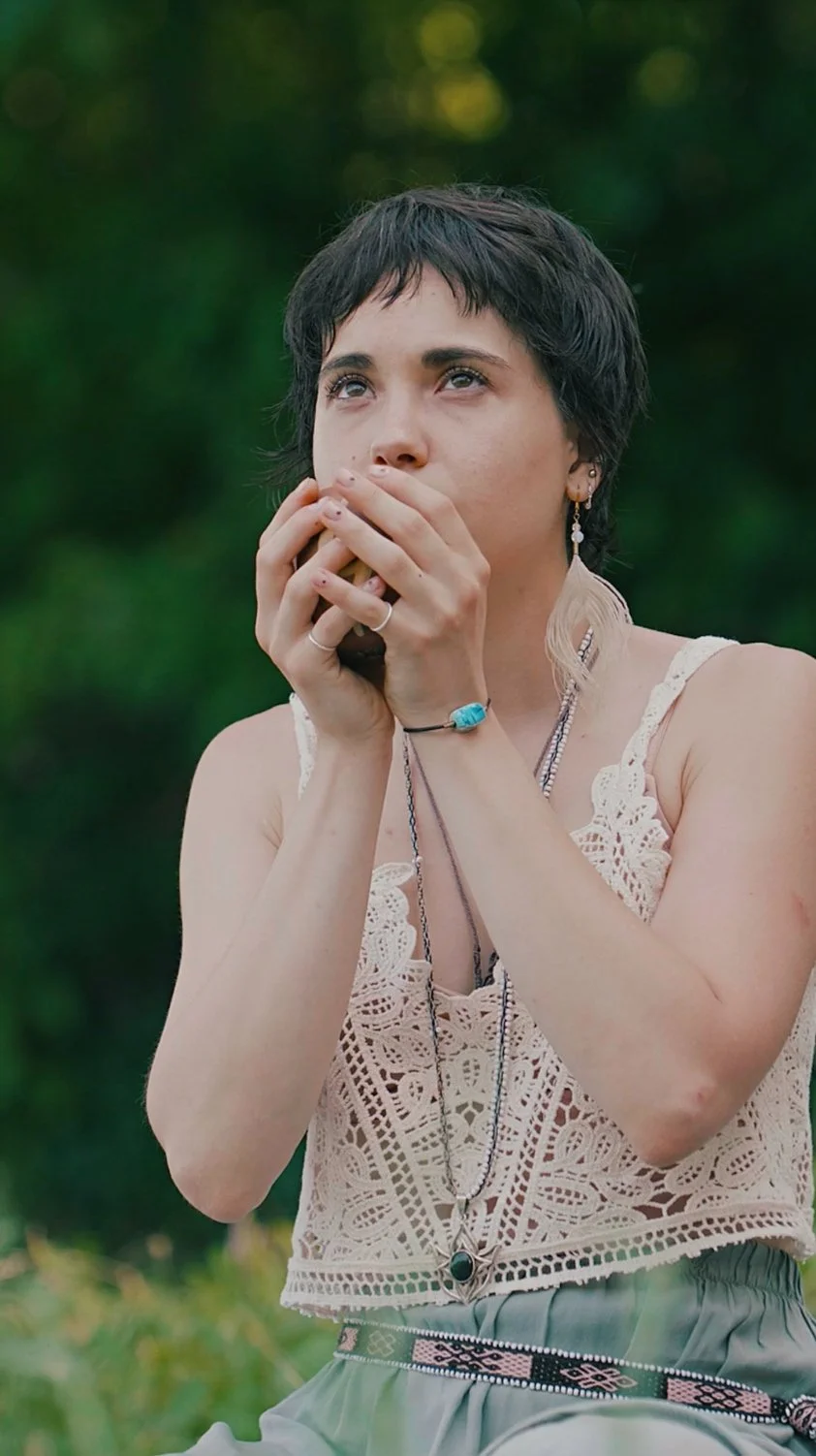 Ragazza con cappello che fuma un sigaro, indossa top in pizzo e accessori etnici, sfondo naturale verde.