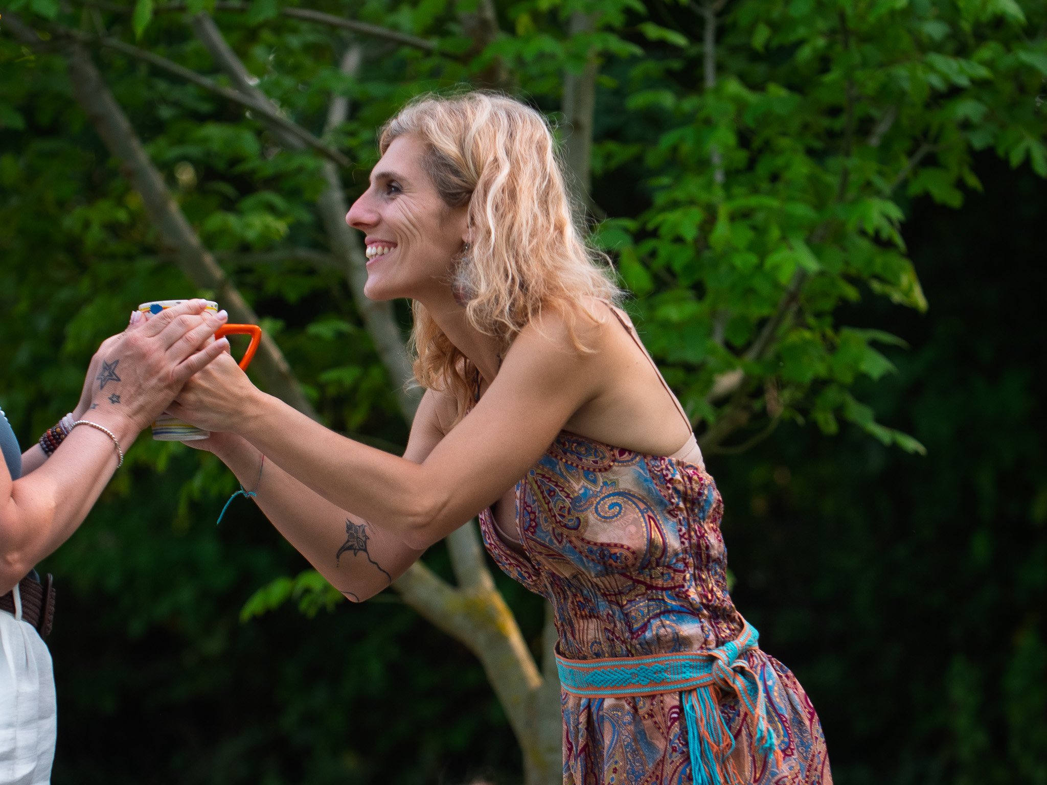 Donna sorridente che riceve una tazza da una persona non visibile in un ambiente naturale con alberi