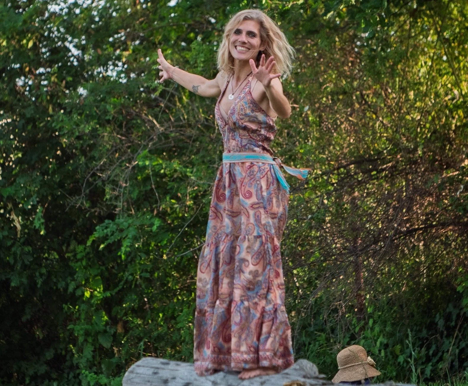 Una donna sorridente in abito lungo con stampa colorata si trova su un parallelepipedo di pietra di fronte a una folta vegetazione, con un cappello di paglia ai suoi piedi e le mani aperte verso l'obiettivo.