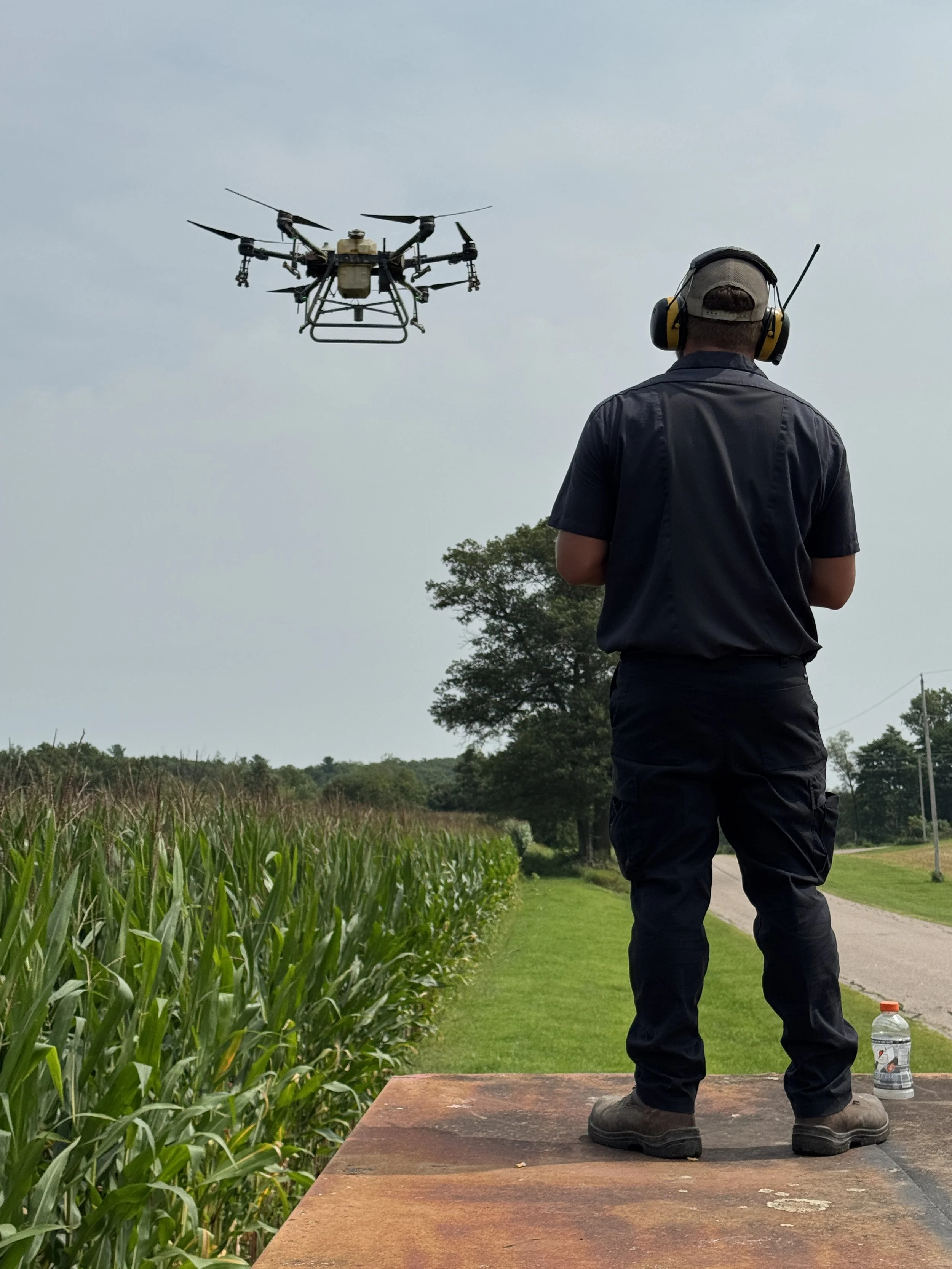 A man in work clothes with headphones and a cap is standing on a platform outdoors, operating or observing a drone flying in the sky over a farm field with corn crops and a tree-lined horizon.