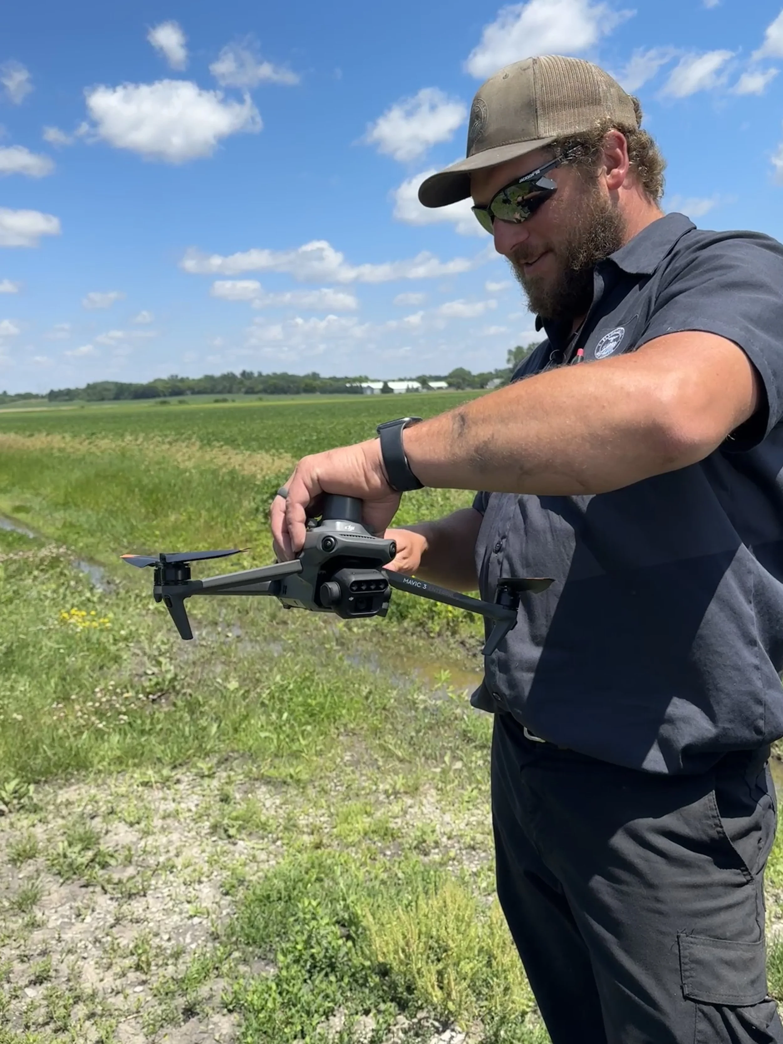 A man wearing sunglasses, a cap, and a dark shirt is holding and preparing a drone in a grassy field with a farm in the background and a blue sky with scattered clouds.