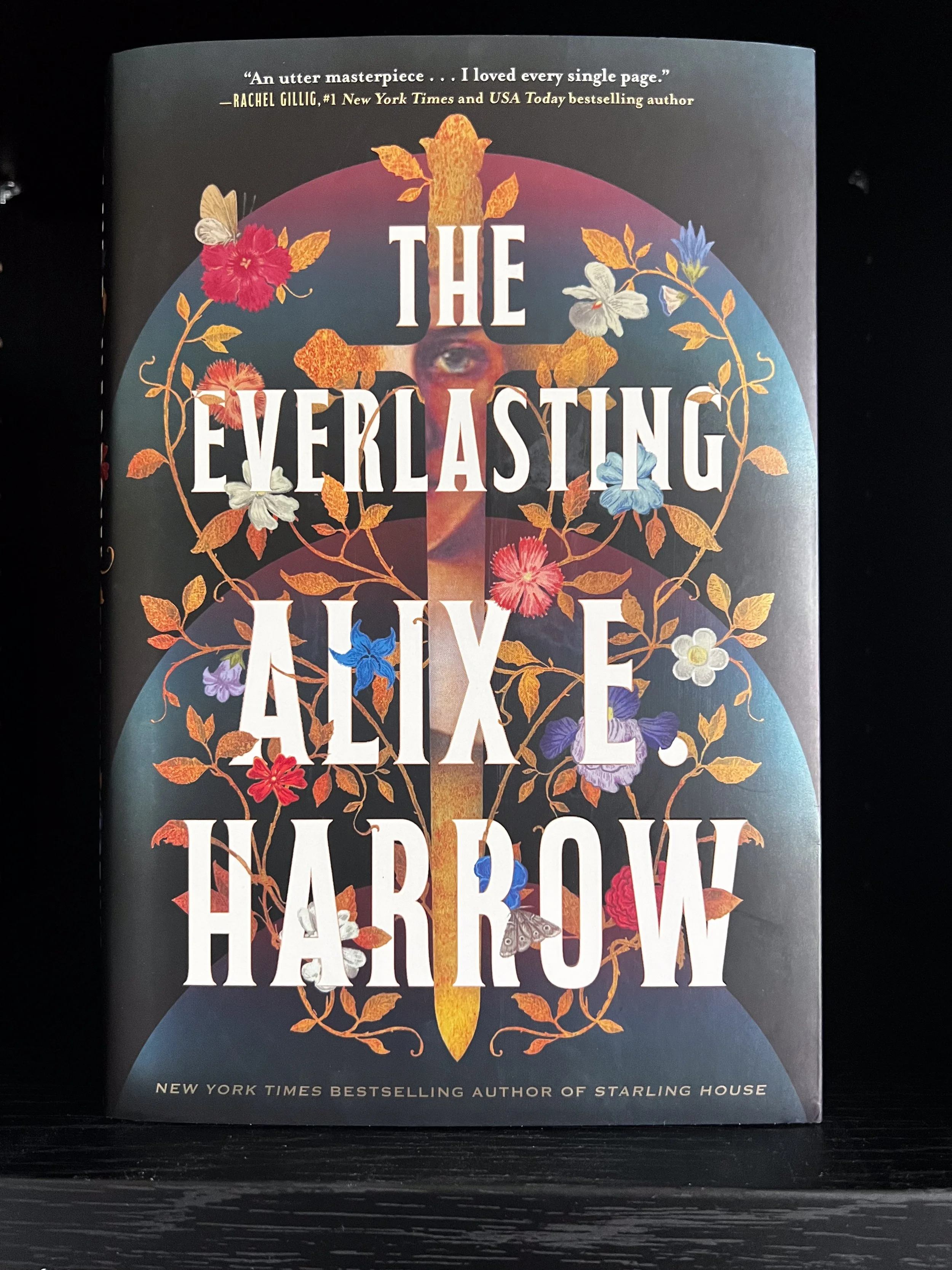 This is the cover of the deluxe edition of The Everlasting by Alix E. Harrow with flowers and a sword with an eye on the hilt of the blade.