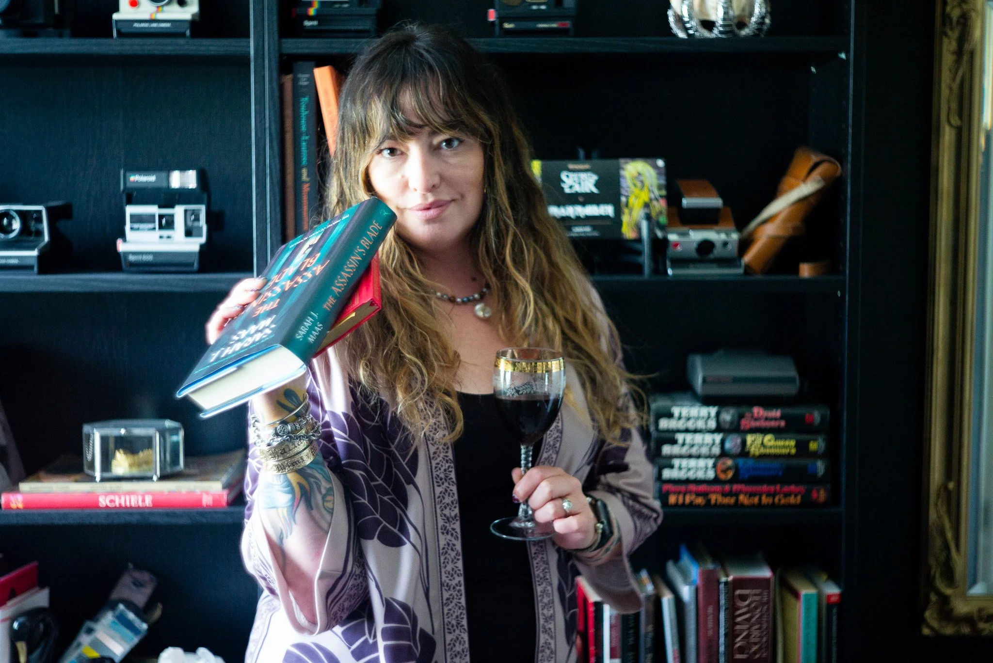 Cassandra is standing in front of black book cases with bold fantasy books shelved behind.  She holds a glass of red wine in one hand and a stack of fantasy and books in another.