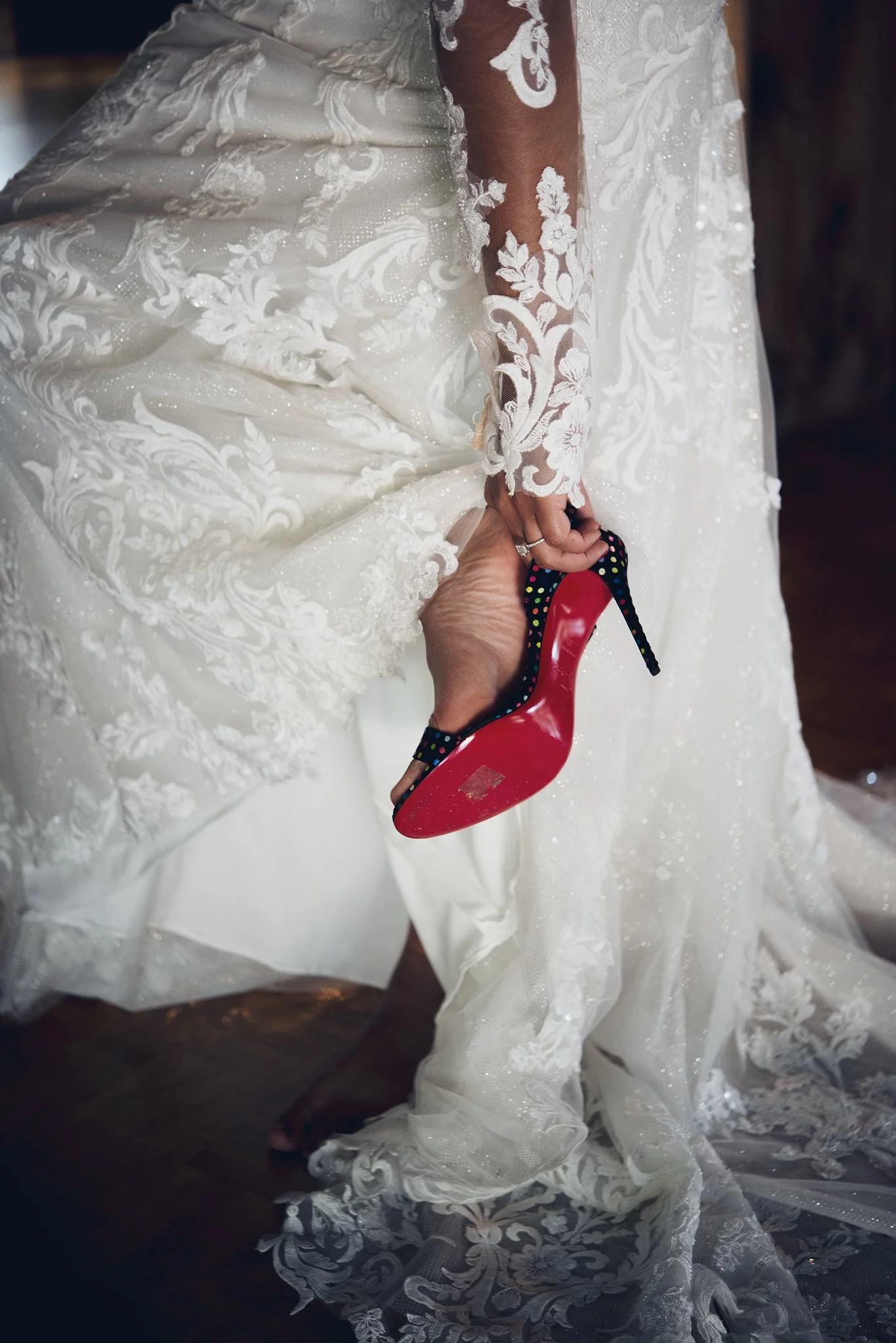 A bride in a white wedding dress with floral lace and embroidery, holding a red high-heeled shoe with black and colorful polka dots on the heel.