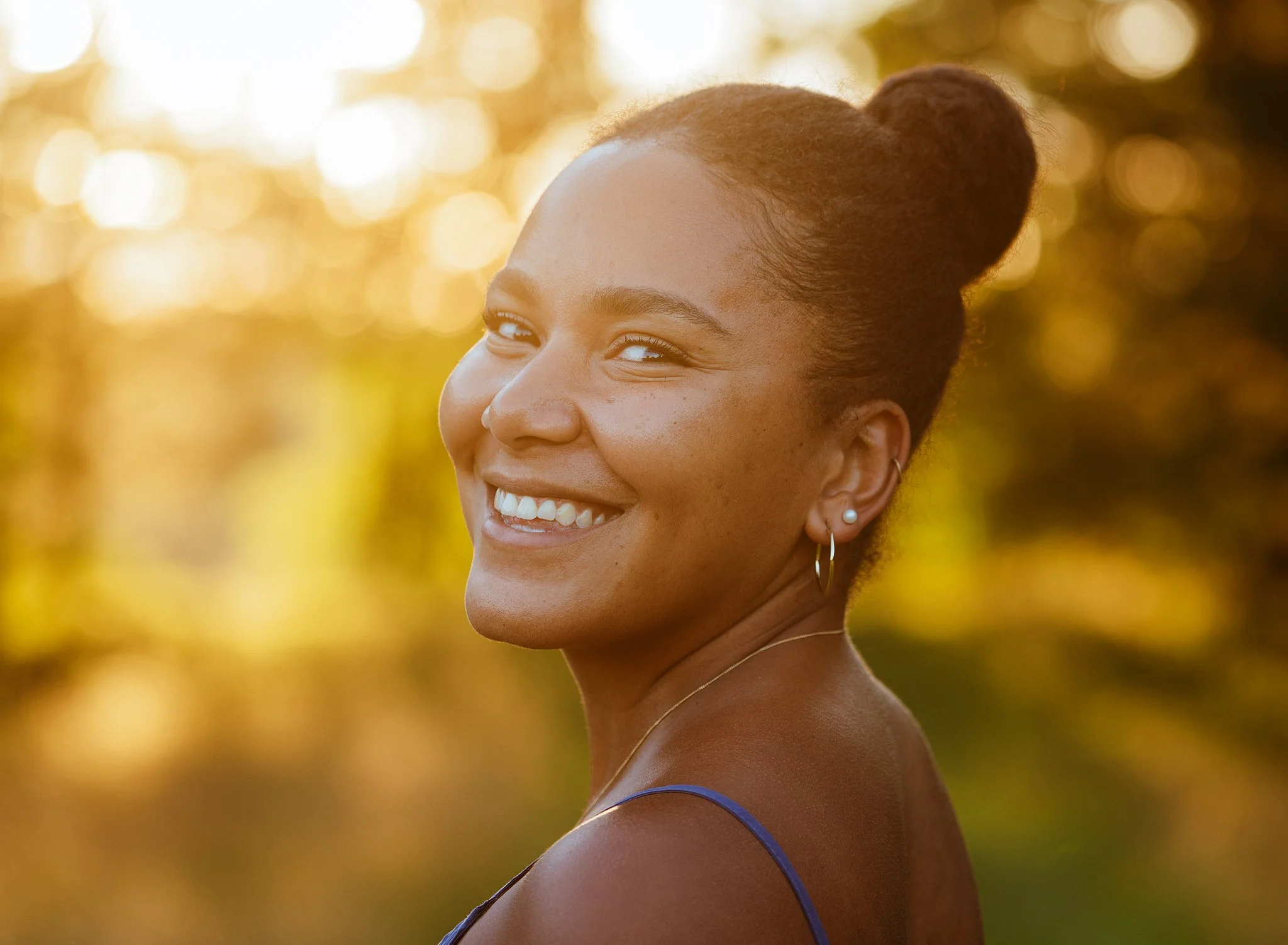 A close-up profile of a woman smiling with white teeth, wearing earrings and a thin necklace, outdoors during golden hour with blurred trees and sunlight in the background.