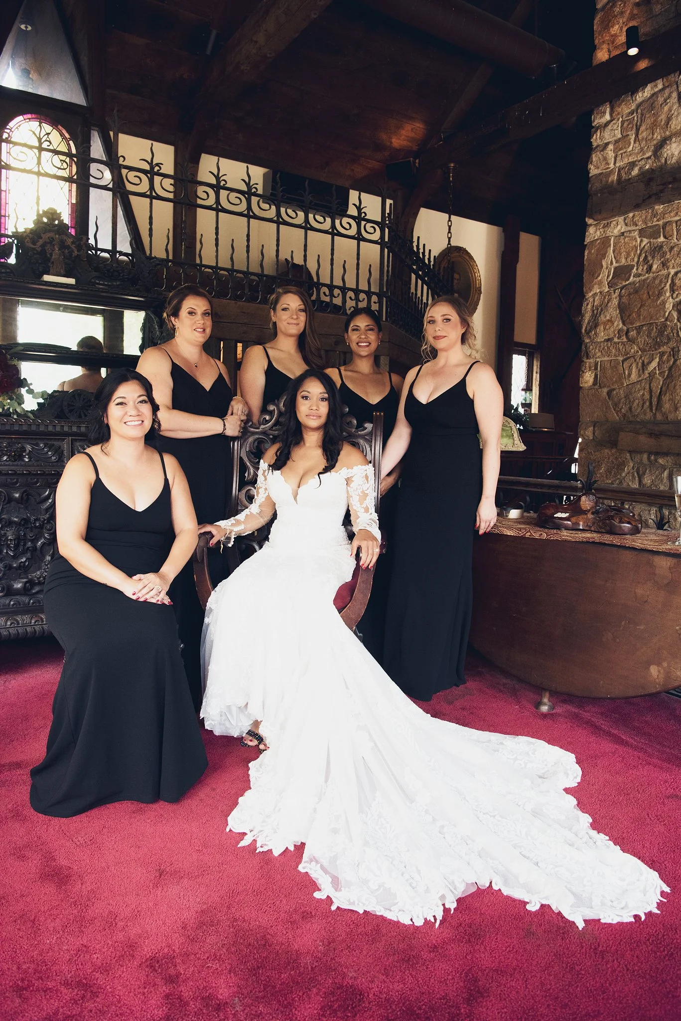 Group of six women, including a bride in a white wedding gown, posing inside a cozy, rustic room with stone and wood walls, stained glass window, and red carpet.