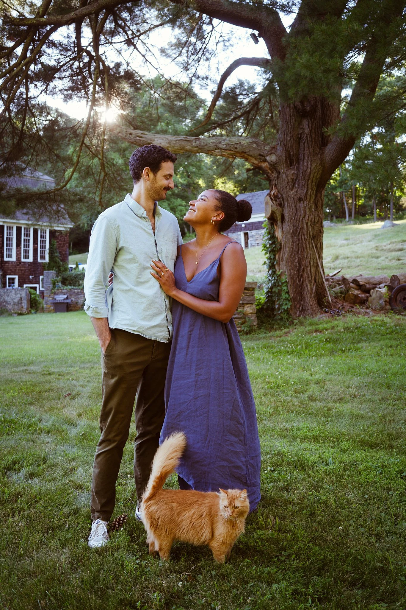 A man and woman standing in a grassy yard under a tree, smiling and looking at each other. A large, fluffy, orange cat stands on the grass near them, and the sun is shining through the branches of the tree.
