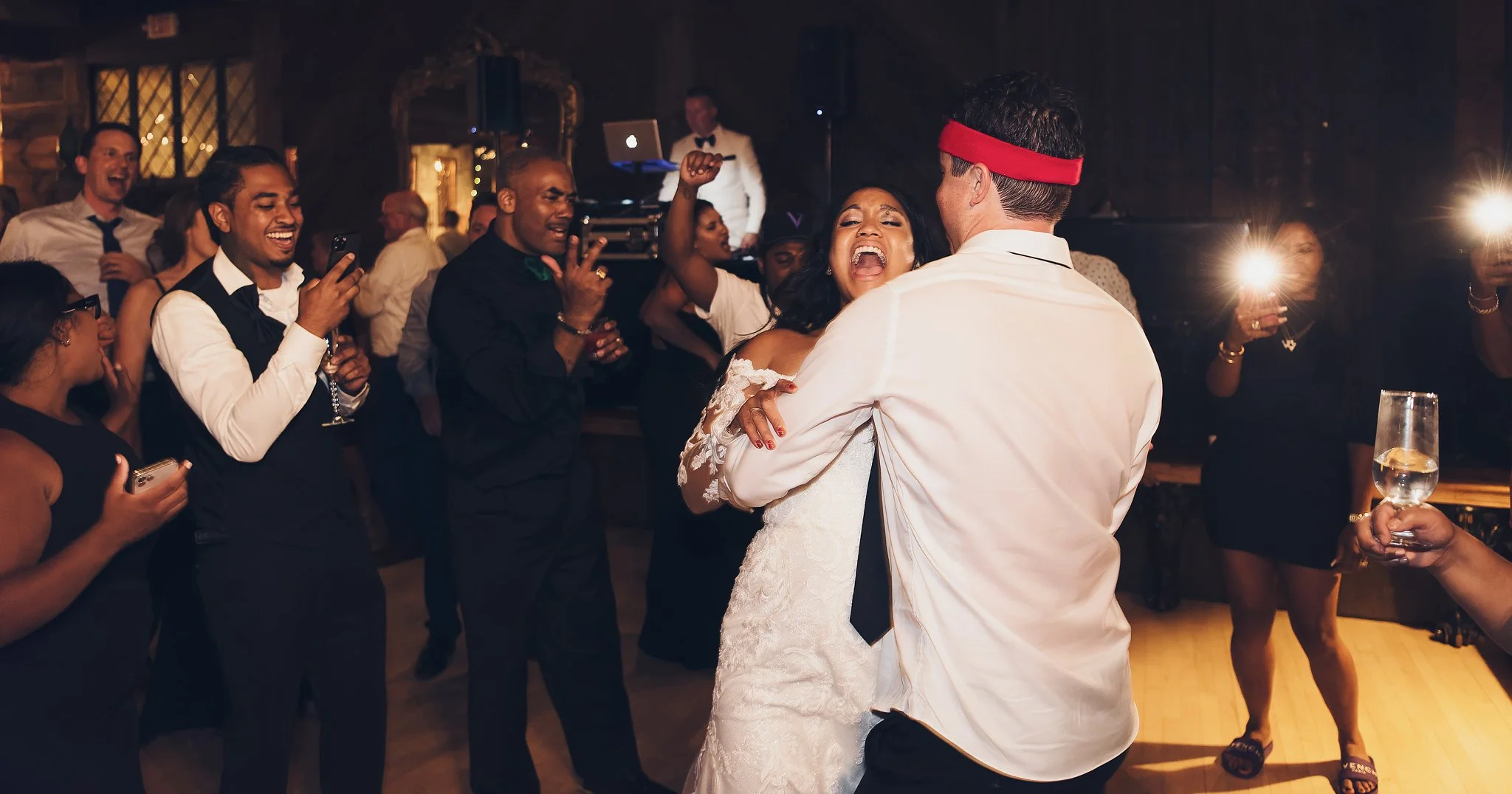 People dancing and celebrating at a wedding reception, with a couple embracing on the dance floor, surrounded by friends taking photos and enjoying the moment.