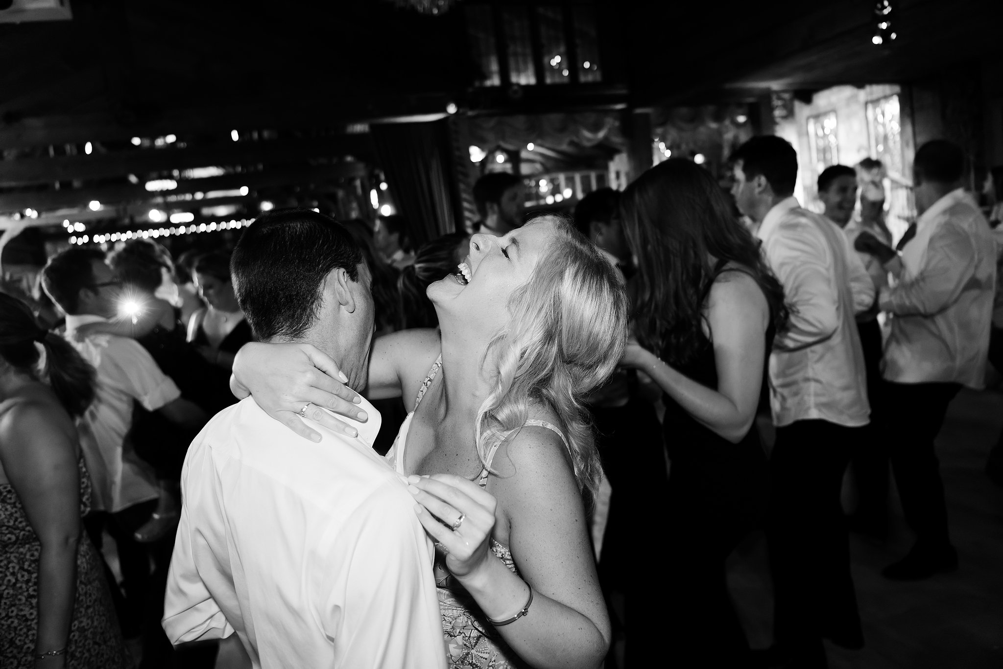 A couple is dancing together at a crowded indoor event, smiling and looking happy. The woman has long wavy hair, and the man is wearing glasses. The background shows other guests engaged in conversations and dancing.