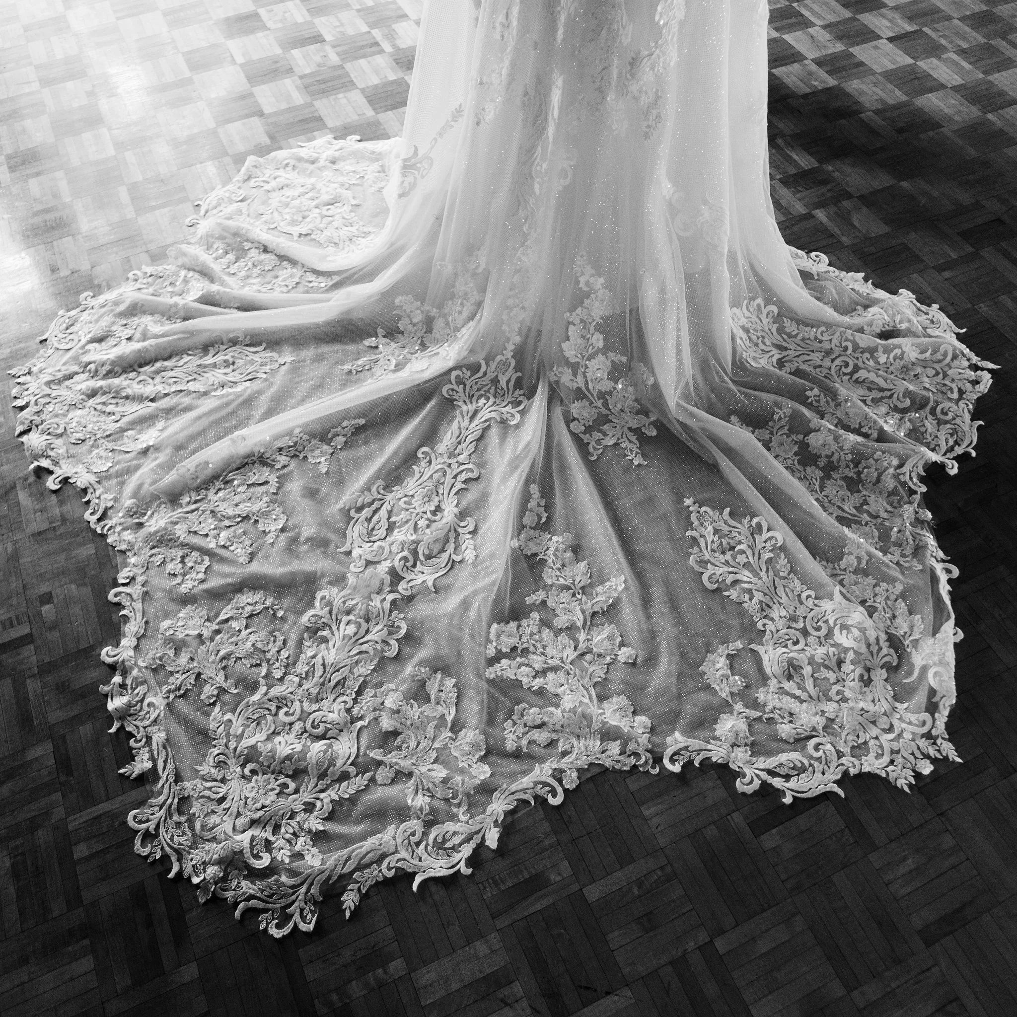Floor length wedding dress with lace detailing and a long train, displayed on a black wooden floor.