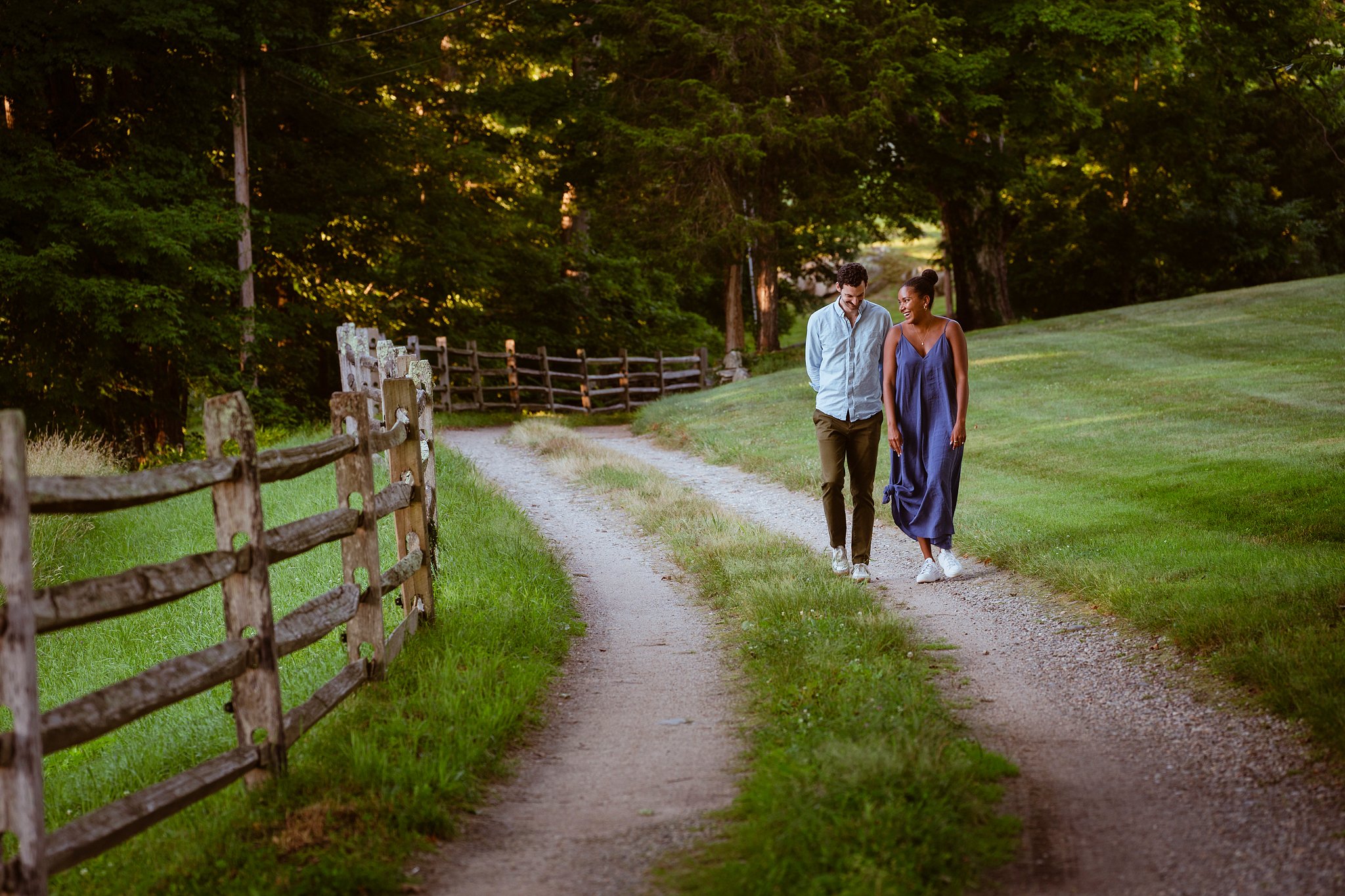 A man and a woman walk along a dirt path in a lush green park, surrounded by tall trees and a wooden fence, during daylight.
