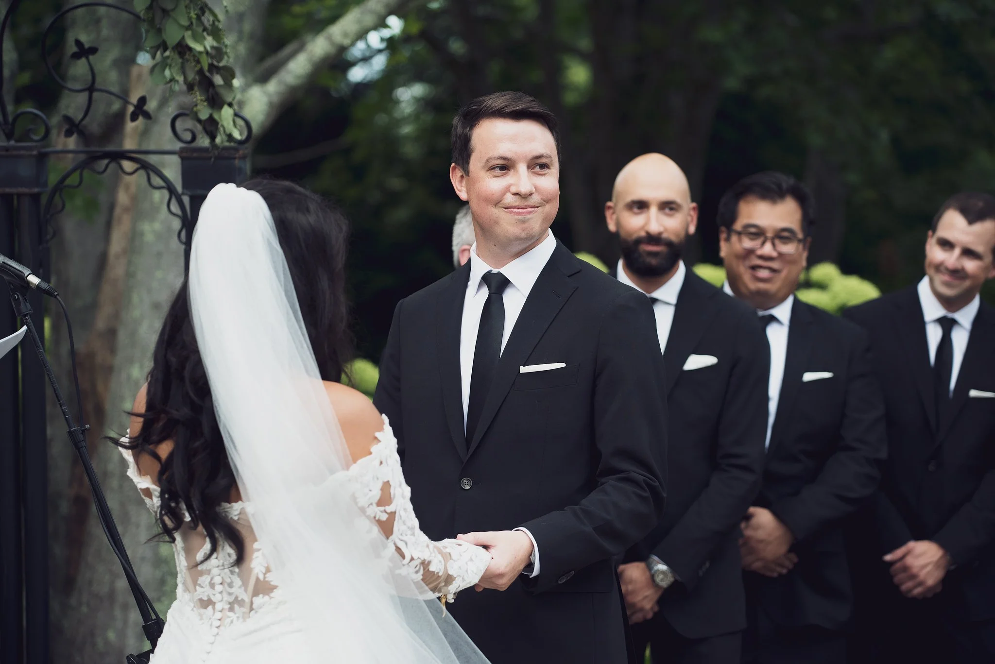 A groom in a black suit and tie holding hands with a bride in a white wedding dress and veil during an outdoor wedding ceremony