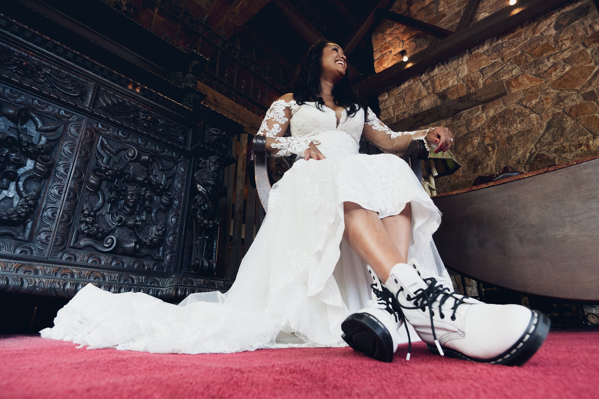 A woman in a wedding dress sitting on a chair, smiling, with her legs crossed, wearing sneakers, indoors with a brick wall and wood ceiling.
