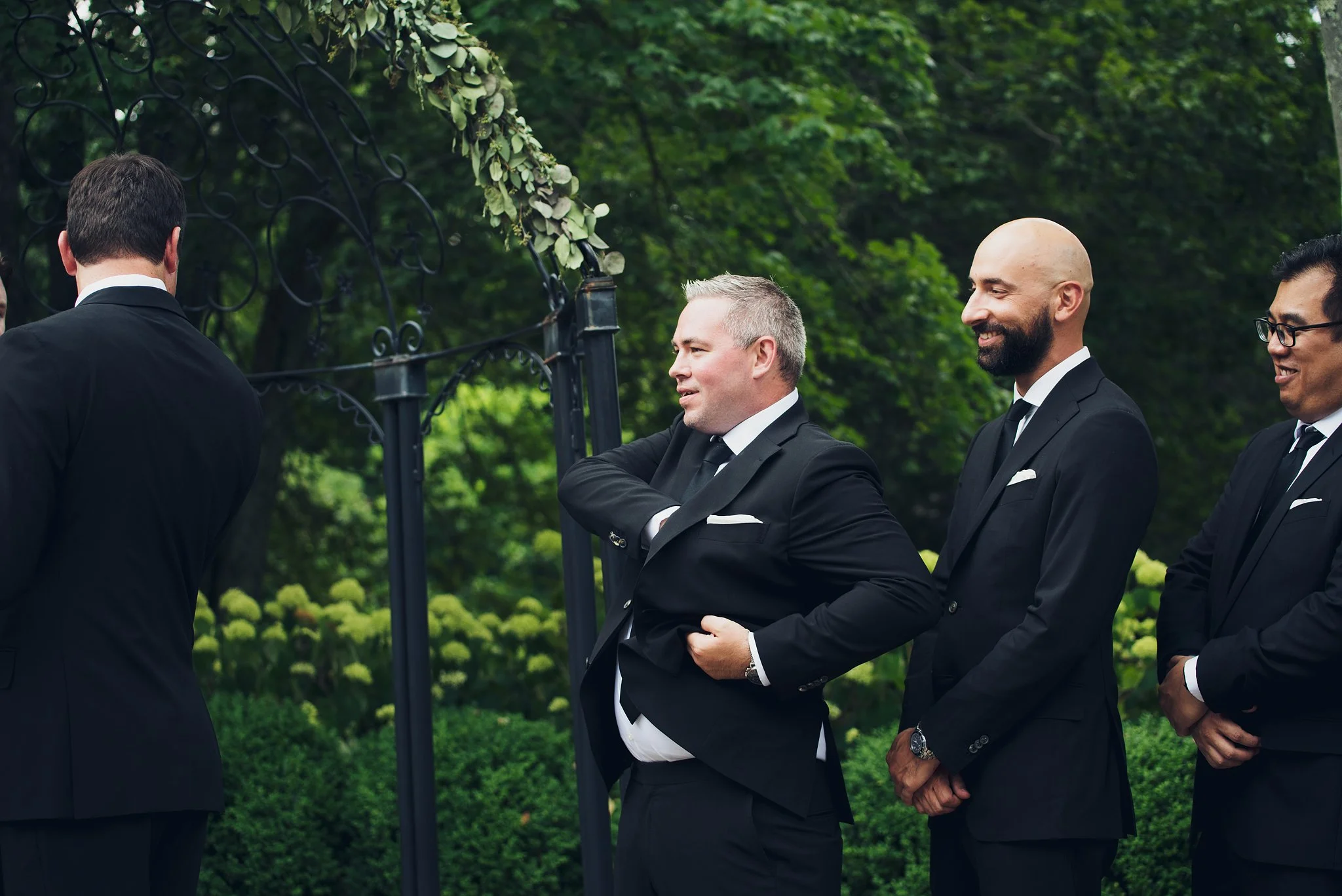 Group of men dressed in black suits and ties standing outdoors, participating in a formal event or ceremony surrounded by greenery.
