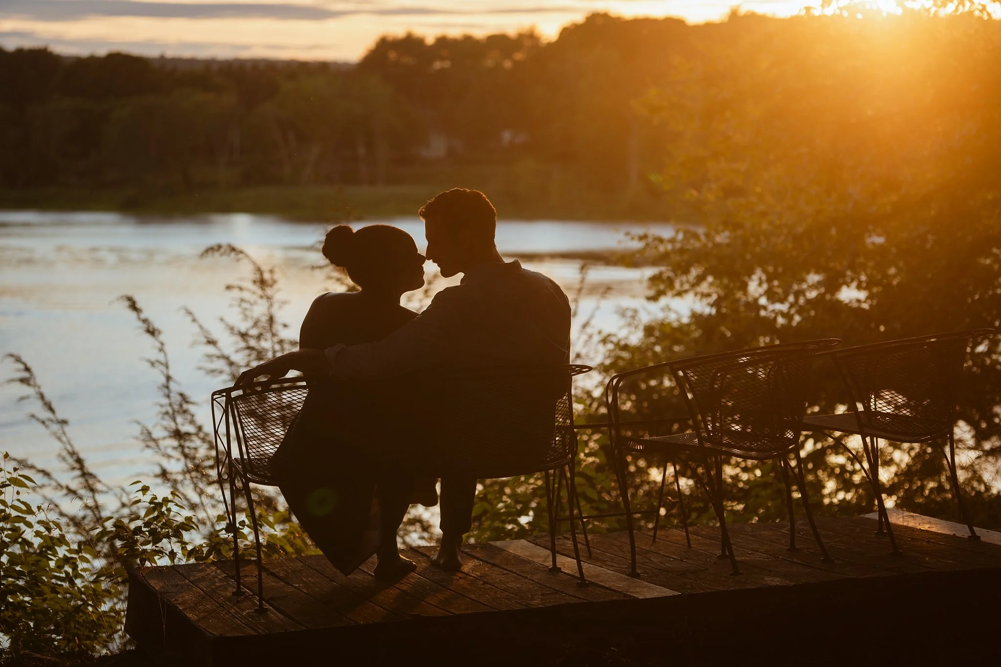 Silhouettes of a couple sitting close together on a porch overlooking a body of water during sunset, with trees and foliage in the background.