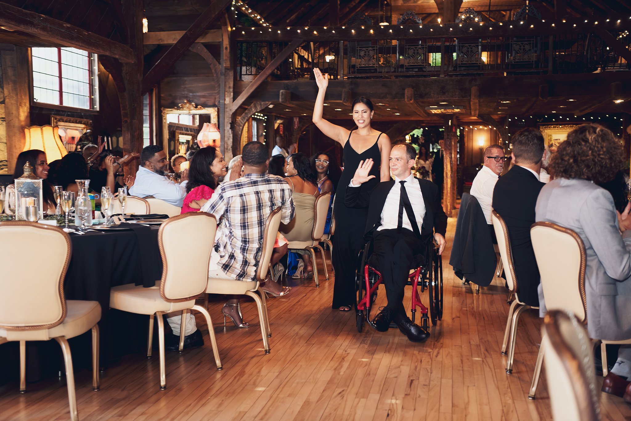 A diverse group of people are seated around tables in a warmly lit rustic restaurant, with some clapping and smiling while a woman in a wheelchair waves and smiles.