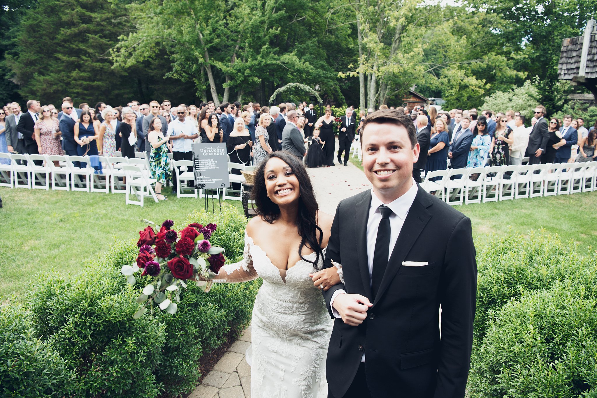 A newlywed couple smiling, walking arm in arm down a garden aisle, with a large crowd of guests standing in the background during an outdoor wedding ceremony surrounded by green trees.