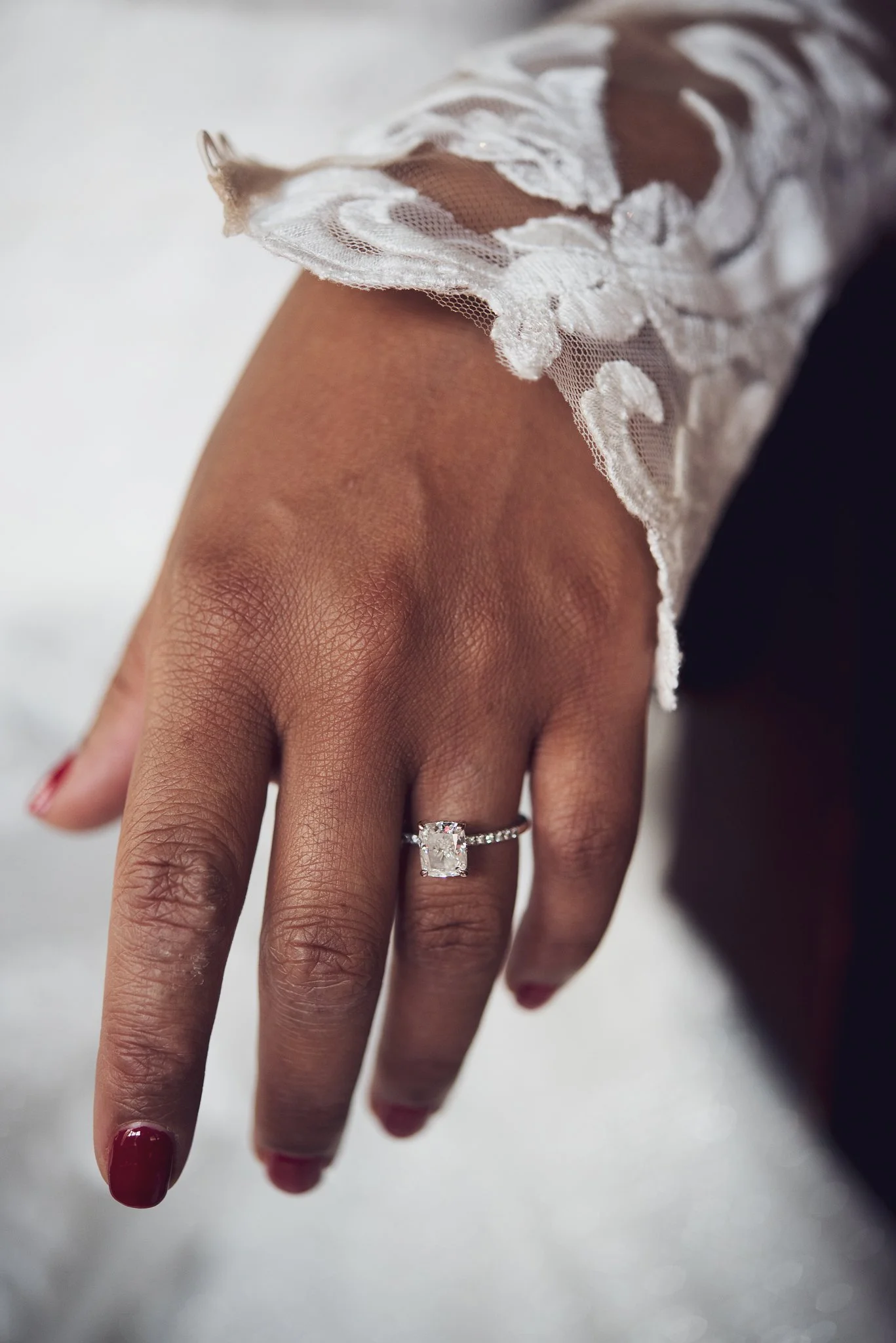 Close-up of a woman's hand wearing a large, square-cut diamond engagement ring with smaller diamonds on the band, resting on a white surface and adorned with a lace sleeve.