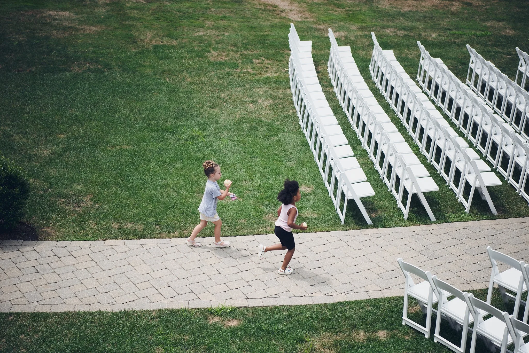 Three children running along a paved walkway next to a grassy area with white folding chairs arranged in rows.