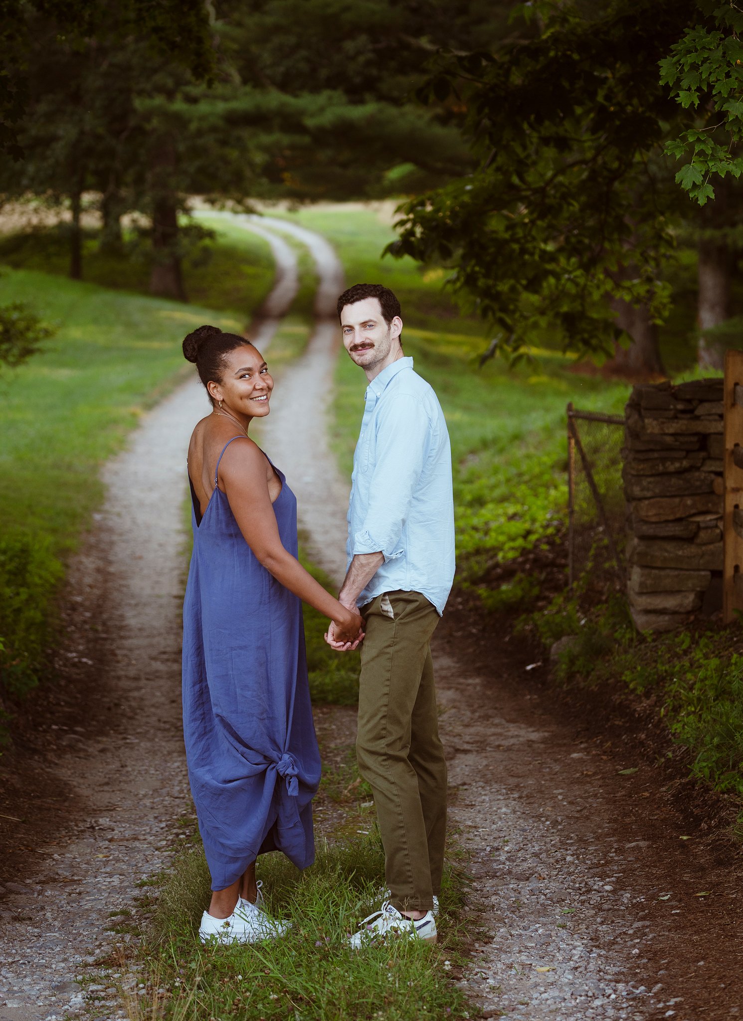 A happy couple holding hands and smiling on a dirt path in a green park with trees and grass.