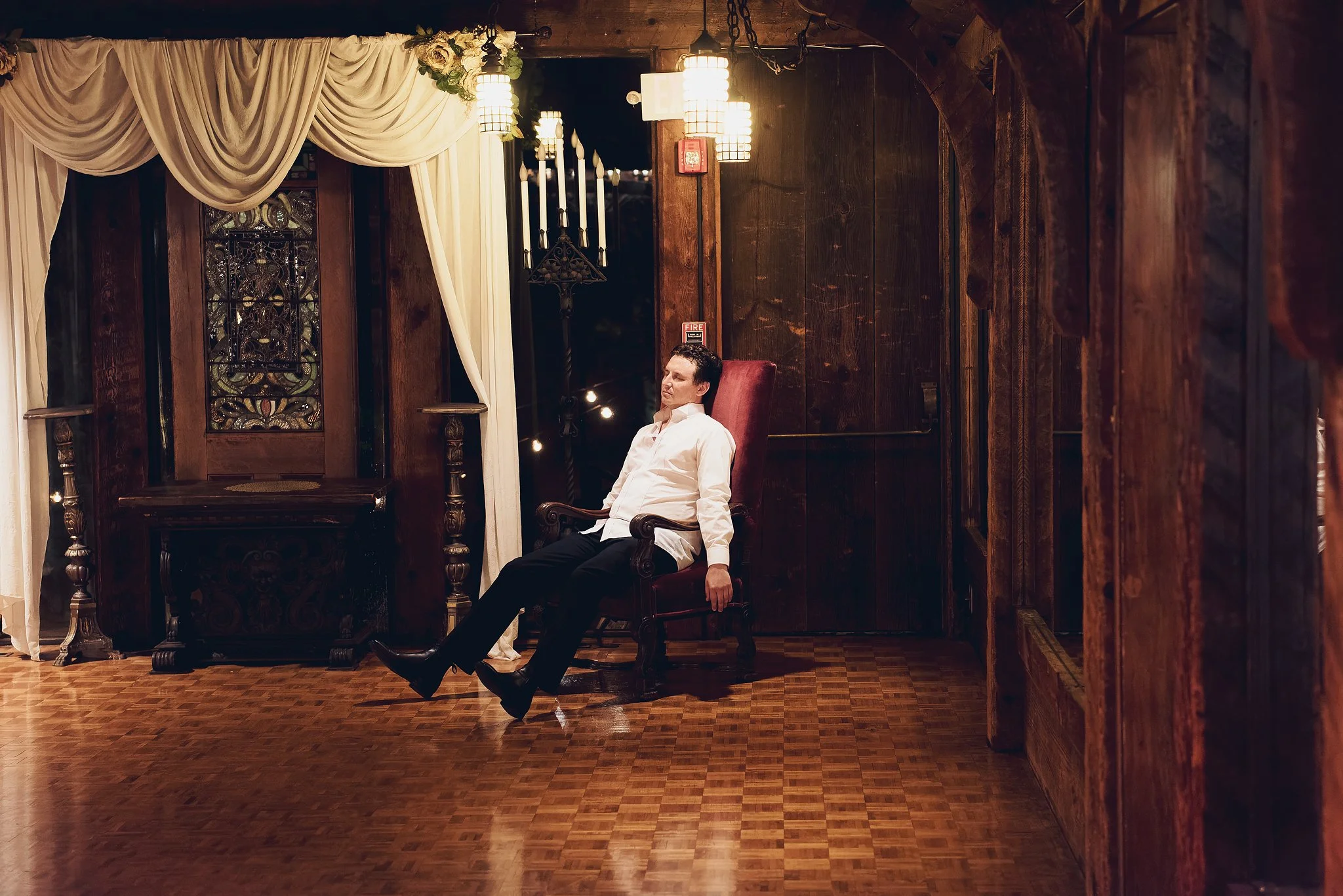 A man seated in a vintage leather armchair in a warmly lit room with dark wood paneling and vintage decor. The room has ornate curtains, stained glass, and hanging lamps, creating a cozy, antique ambiance.