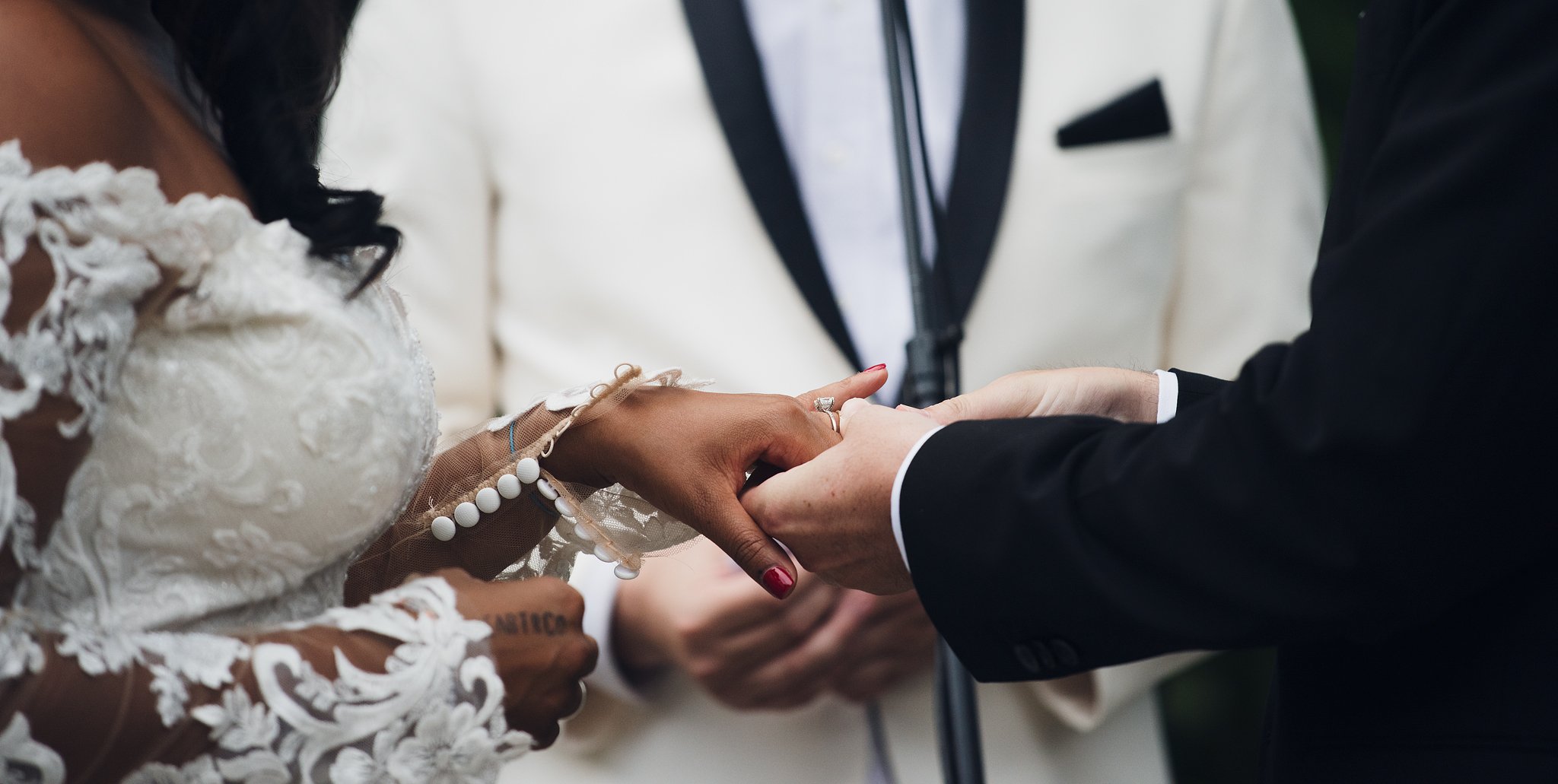 Close-up of a wedding ceremony showing a bride and groom holding hands during the ring exchange. The bride is in a lace wedding dress, and the groom is in a black suit. The focus is on their hands and the rings.