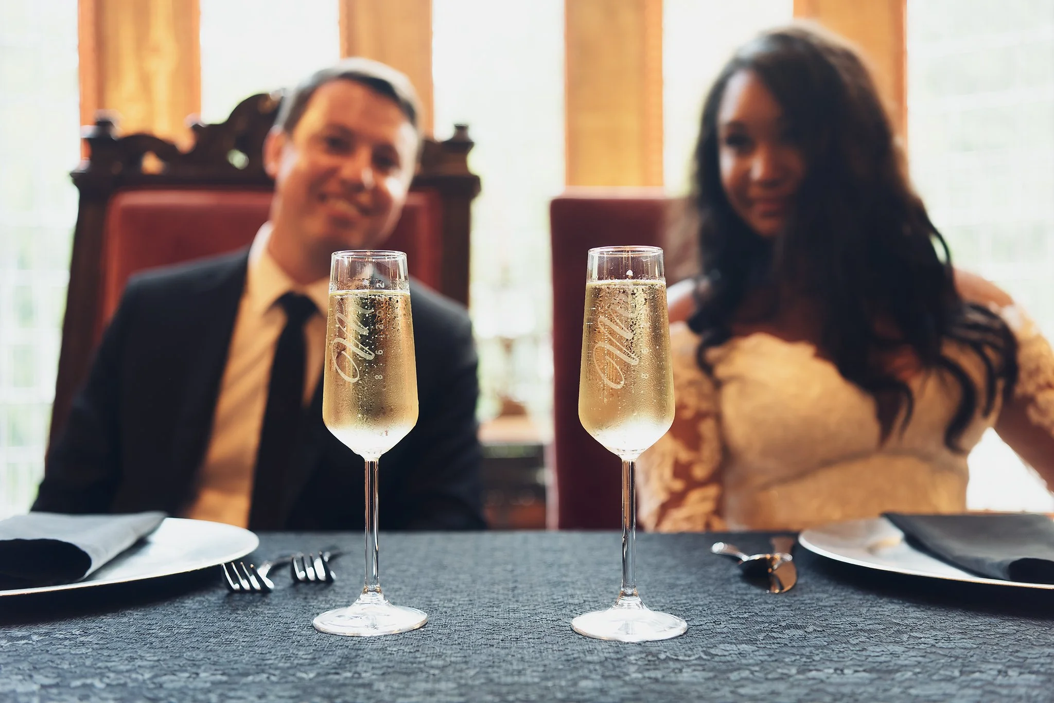 Two glasses of champagne on a dining table set for a formal dinner, with black napkins, plates, forks, knives, and spoons. In the background, a happy couple in formal attire, a man in a suit and tie and a woman in a fancy dress, sit at the table.
