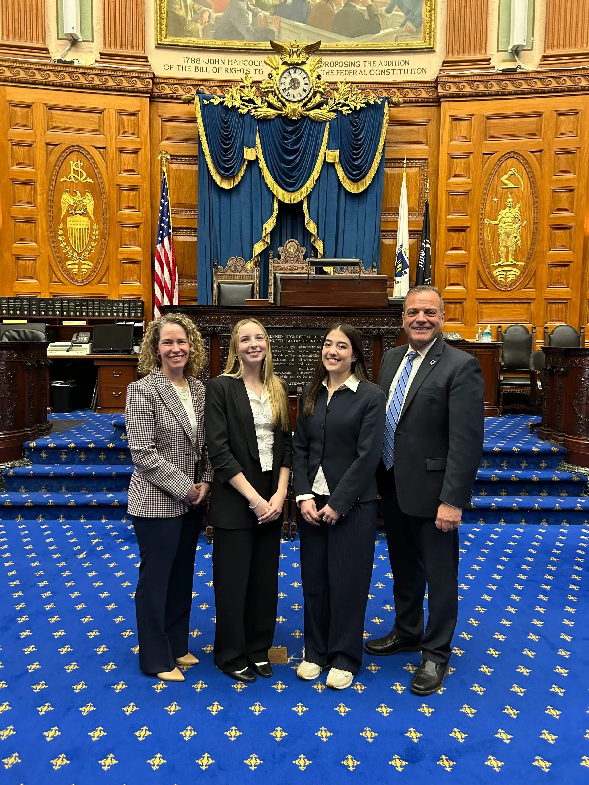 4 people stand in front of a podium of the MA State House