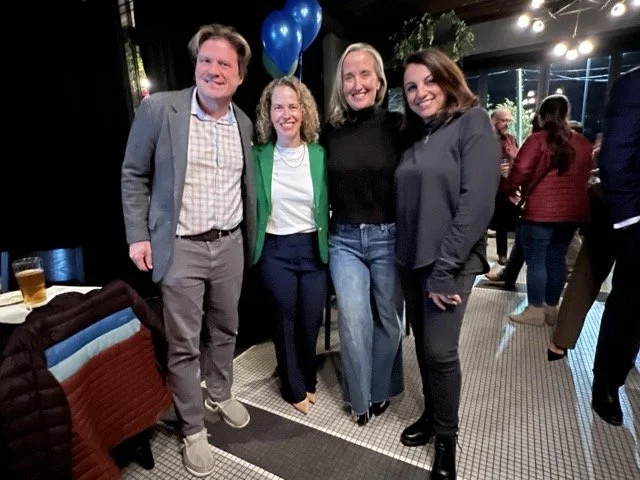 Four school committee members standing together at a social event, smiling at the camera. The group includes two women and two men, with decorations and other attendees in the background.